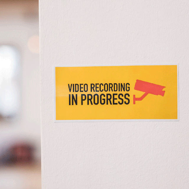 Un panneau jaune sur un mur indique 'VIDEO RECORDING IN PROGRESS' avec une icône de caméra rouge.