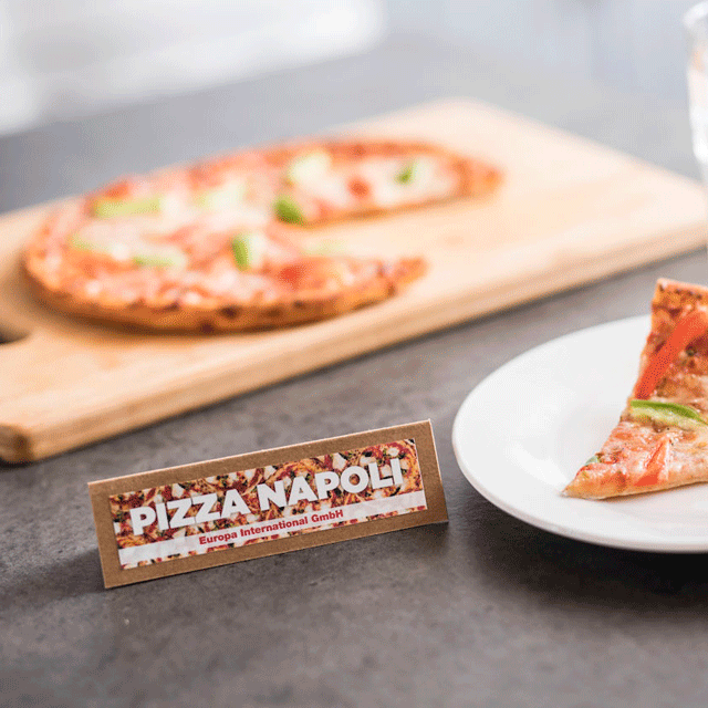 Un panneau 'Pizza Napoli' est près d'une part de pizza dans une assiette et d'une pizza entière sur une planche.