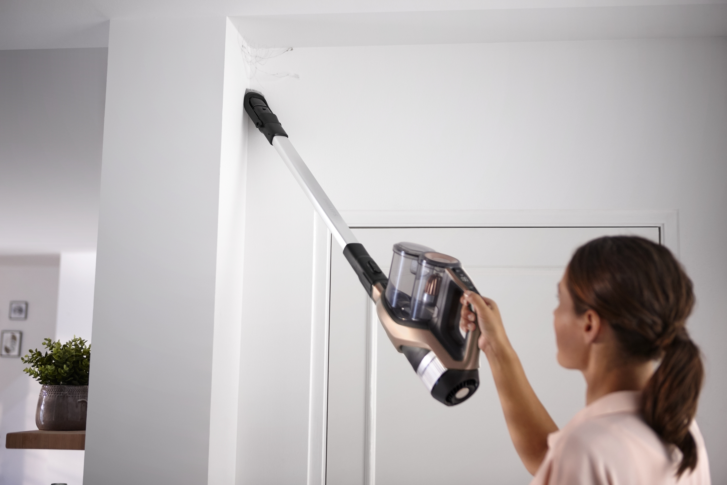 Eine Frau benutzt einen Staubsauger an einer Wand. Der Hintergrund ist eine weiße Wand und eine Tür.