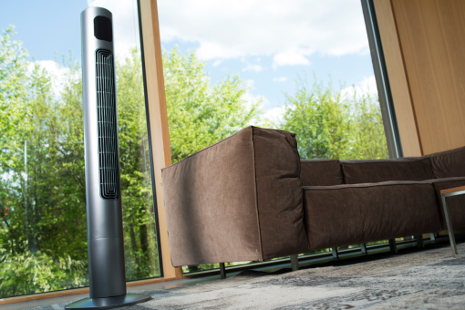 Ein hoher silberner Ventilator steht neben einem braunen Sofa in der Nähe eines großen Fensters mit Blick auf einen grünen Baum.
