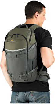 Homme portant un sac à dos vert et gris. Il porte une chemise noire et un jean, dos à la caméra.