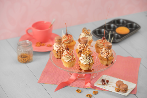 Cupcakes auf einem Glasständer, dekoriert mit Karamell, Schokolade und Brezeln, neben einer rosa Tasse.
