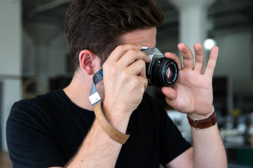 Homme tenant un appareil photo devant son œil, portant une chemise noire et une montre marron.