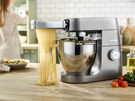 Una macchina da cucina Kenwood argentata sta facendo la pasta. La pasta è appesa su un tavolo di legno.