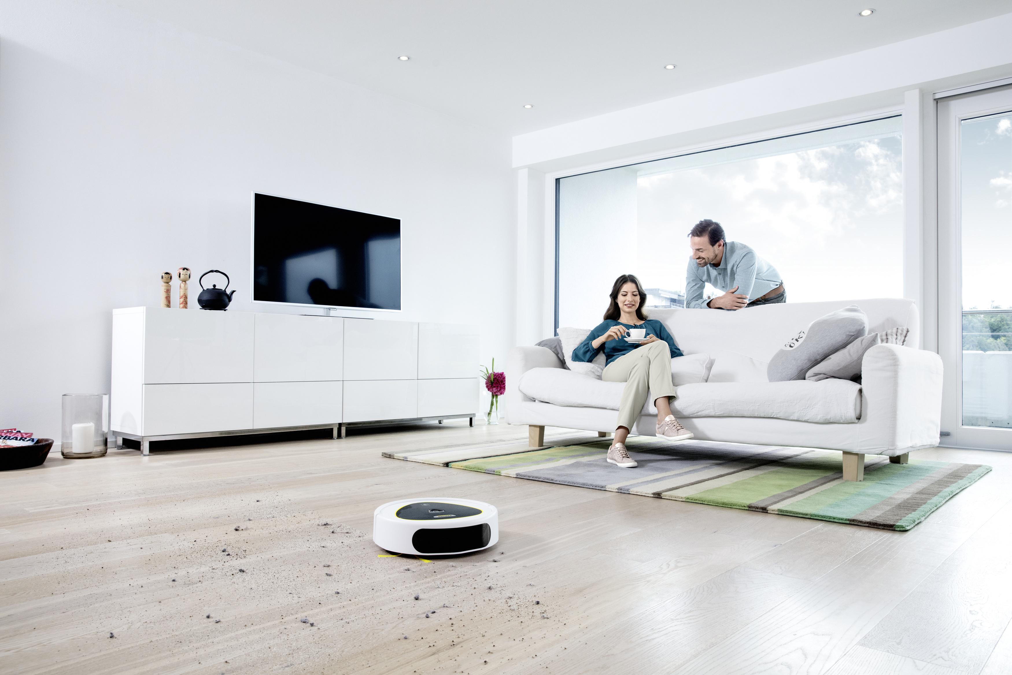 Wohnzimmer mit Paar auf weißem Sofa. Ein Roboter-Staubsauger auf dem Boden. Großes Fenster.