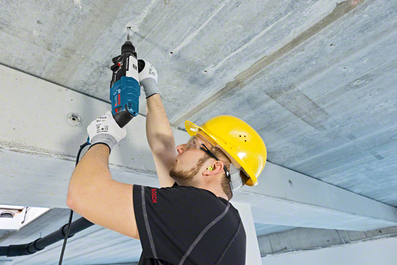Egy férfi sárga védősisakban Bosch fúróval beton mennyezetet fúr.
