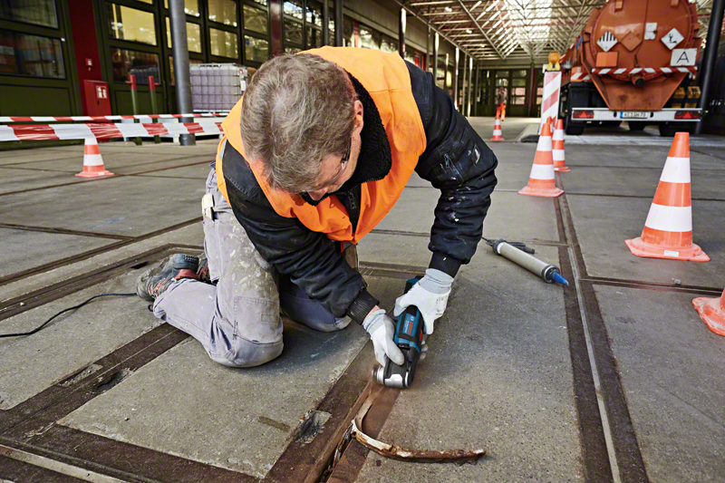 Egy személy hornyot vág a padlóba elektromos szerszámmal, védőfelszerelést viselve.