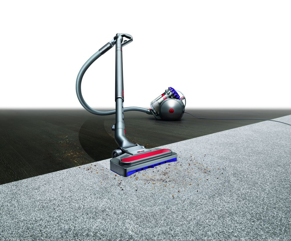 Un aspirateur Dyson gris sur un tapis avec des débris. Arrière-plan : parquet et blanc.