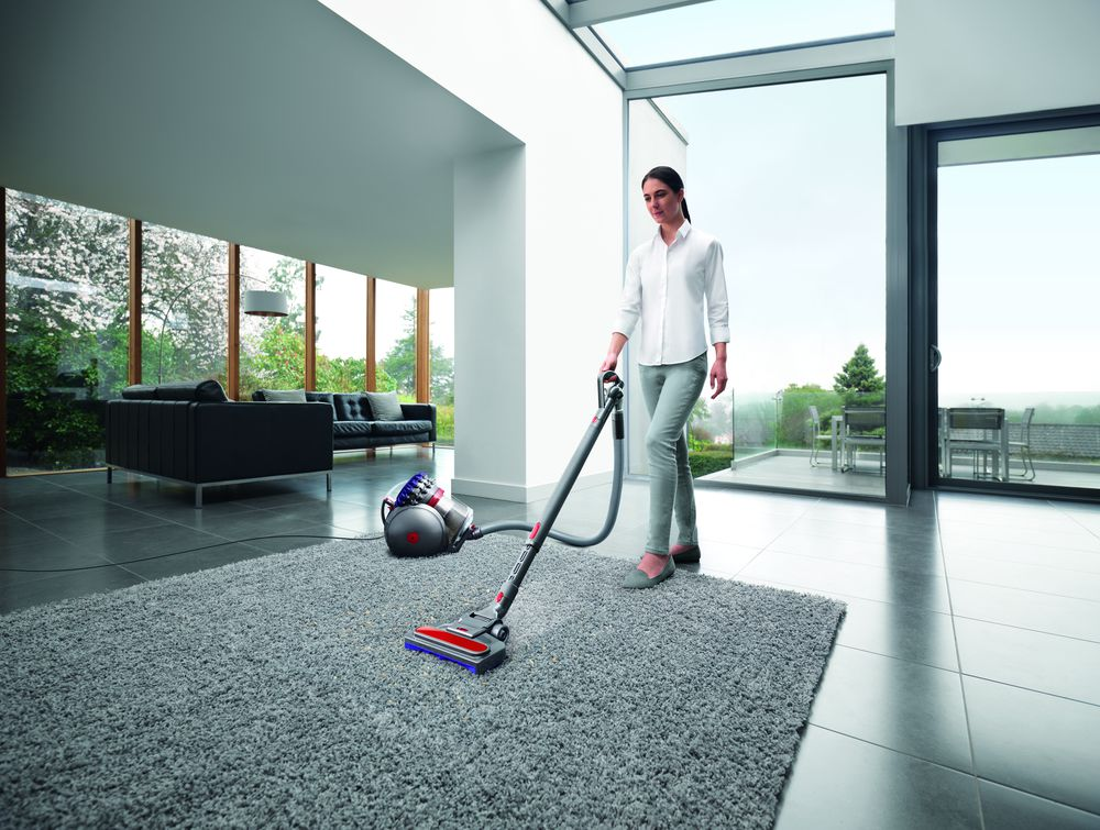 Une femme passe l'aspirateur sur un tapis gris dans un salon moderne avec des baies vitrées.