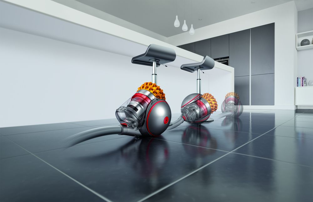 Drie Dyson stofzuigers op een glanzende vloer in een keuken; twee krukken erachter.