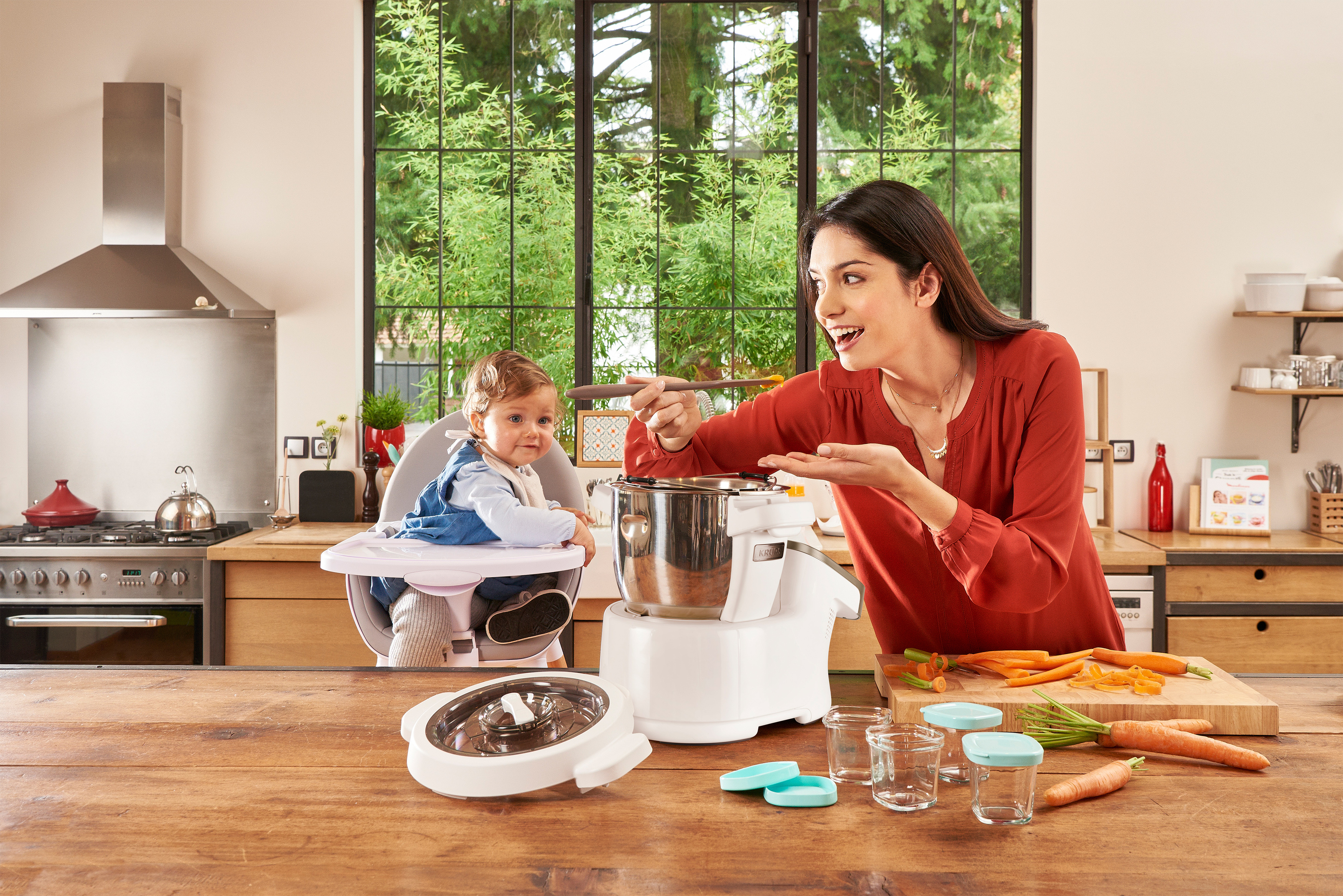 Frau in der Küche mit Baby, Küchenmaschine und Zutaten auf dem Tisch. Sie lächelt und zeigt.