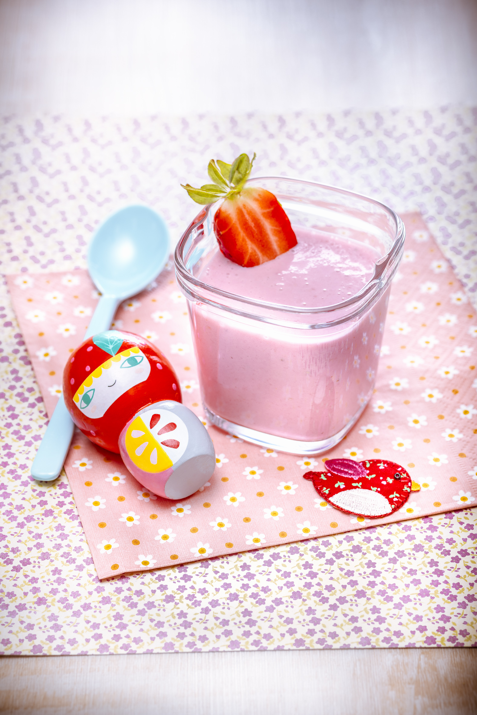 Ein Glas rosa Smoothie mit Erdbeere, Löffel und zwei Spielfiguren auf einer blumenmusternden Serviette.
