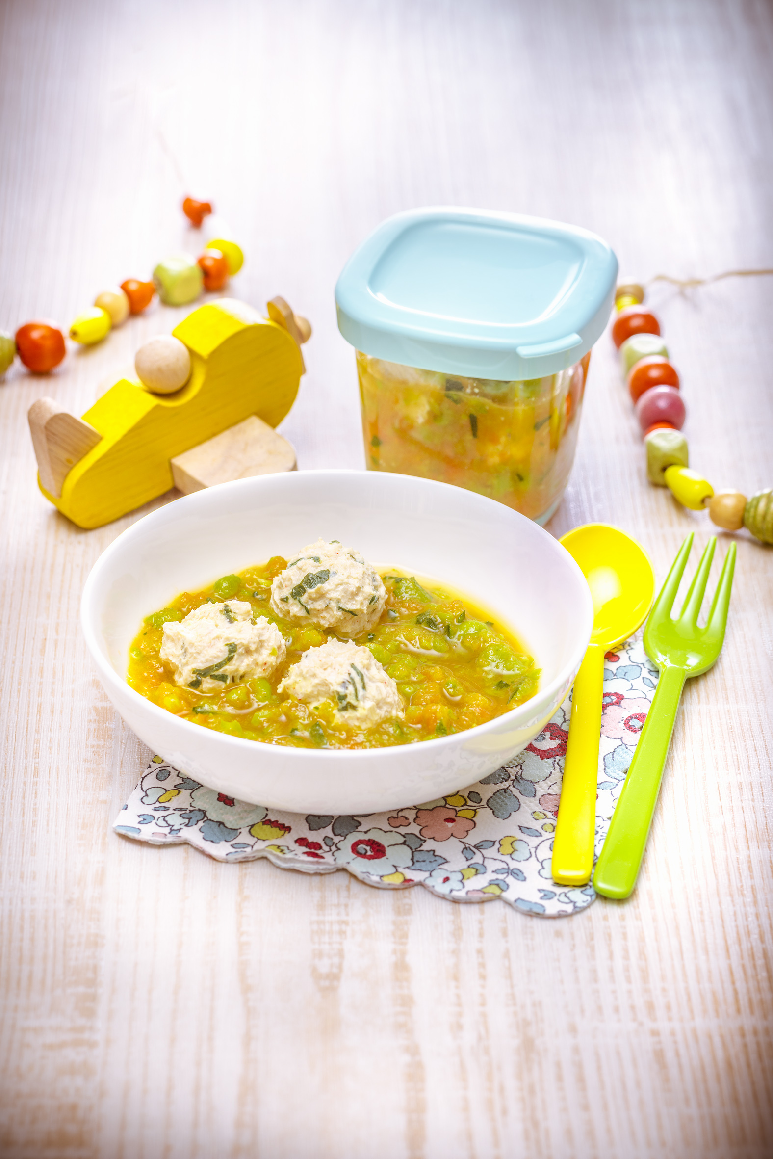 Eine Schüssel mit Fleischbällchen in Sauce, ein Glas Essen, Löffel, Gabel und Spielzeugflugzeug auf einem Tisch.