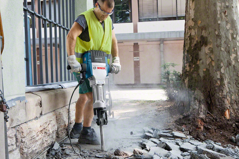 Férfi kalapáccsal töri a betont. Védőszemüveget és fényvisszaverő mellényt visel.