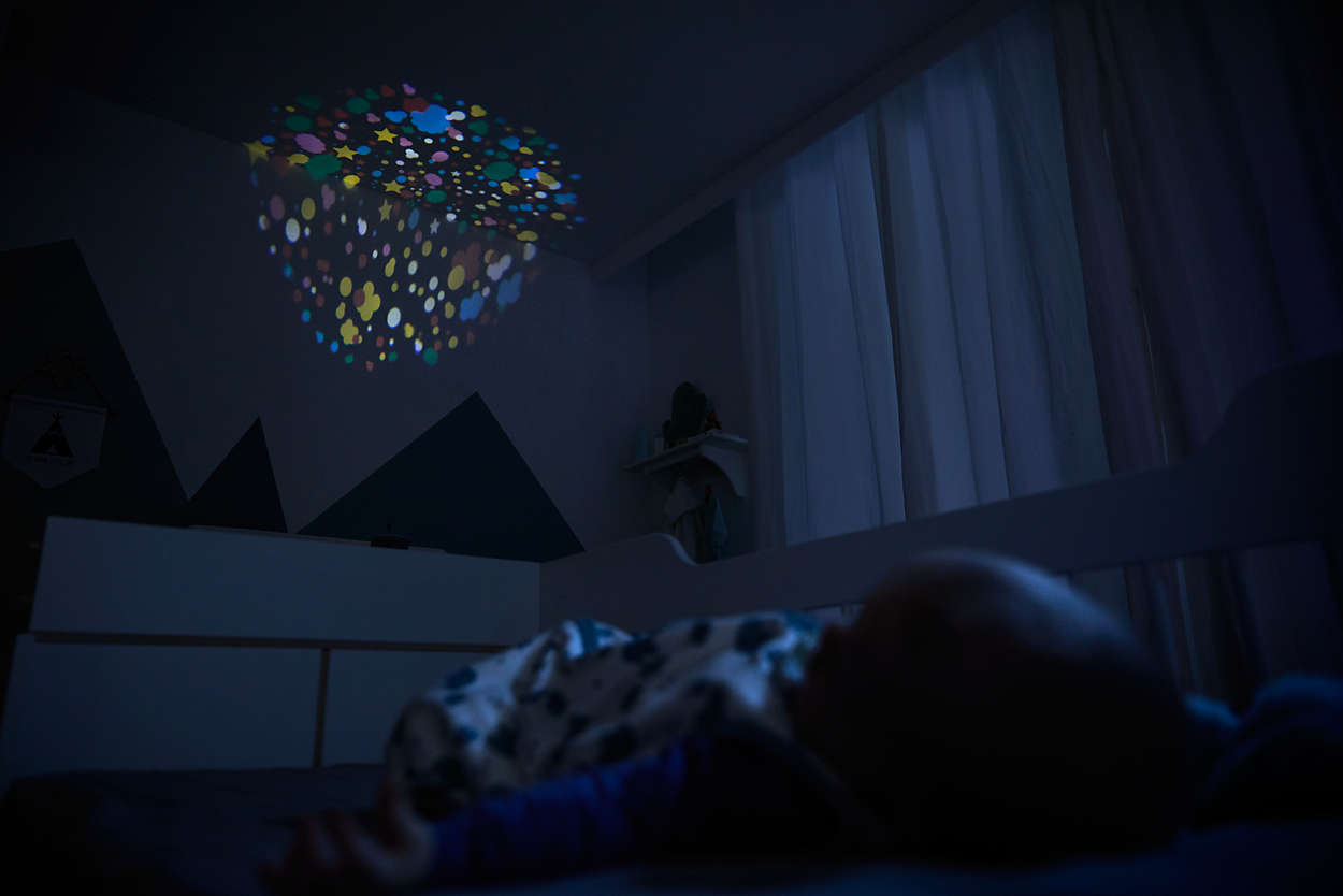 Ein Baby schläft in einem dunklen Schlafzimmer, mit einer farbenfrohen sternförmigen Lichtprojektion an der Decke.