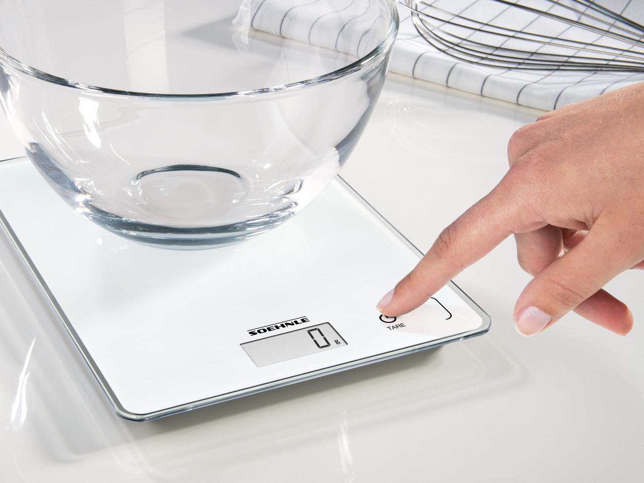 Une main appuie sur un bouton sur une balance blanche. Un bol en verre est sur la balance.