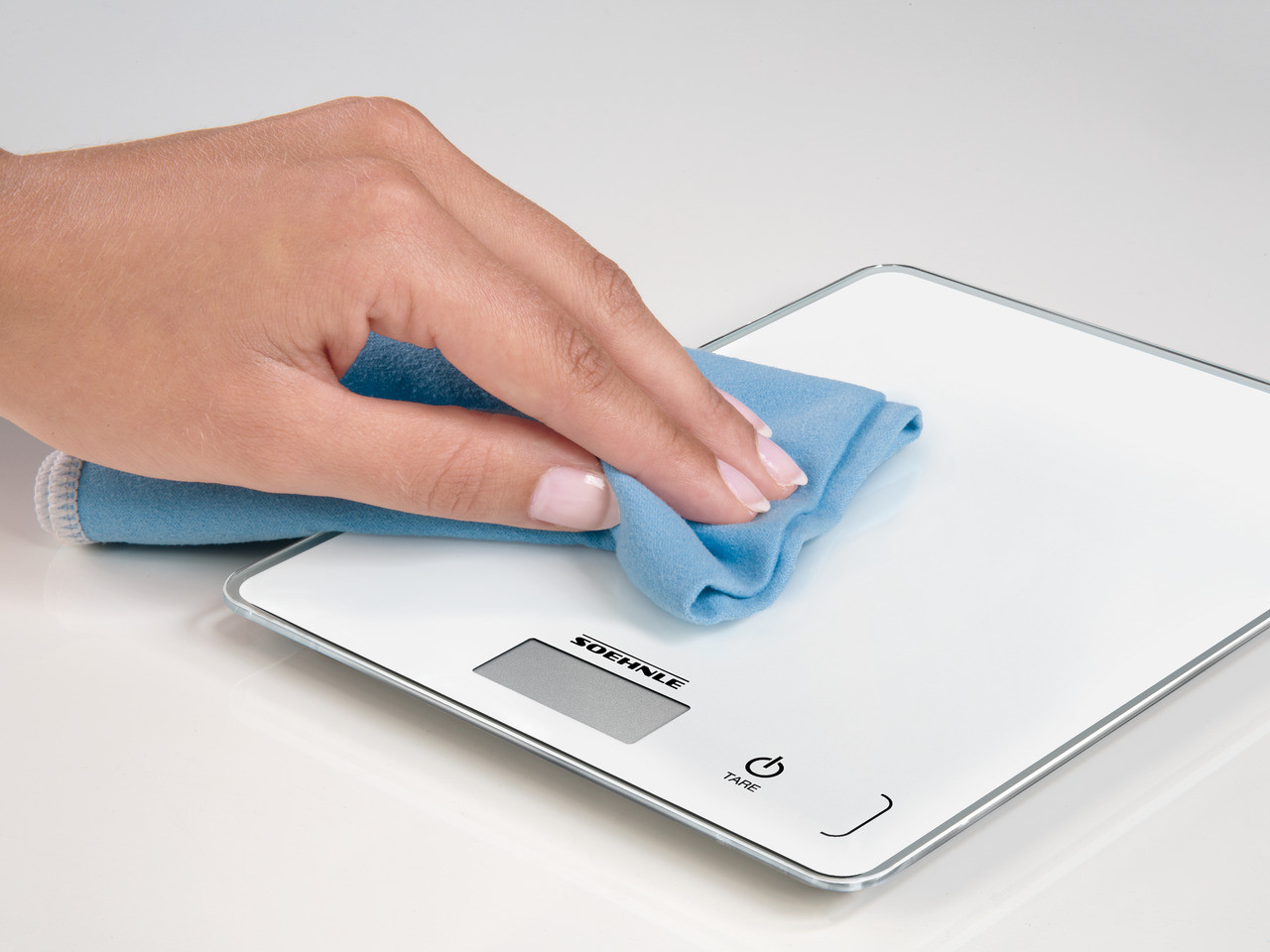 Une main essuie une balance numérique blanche avec un chiffon bleu. Le fond est blanc.