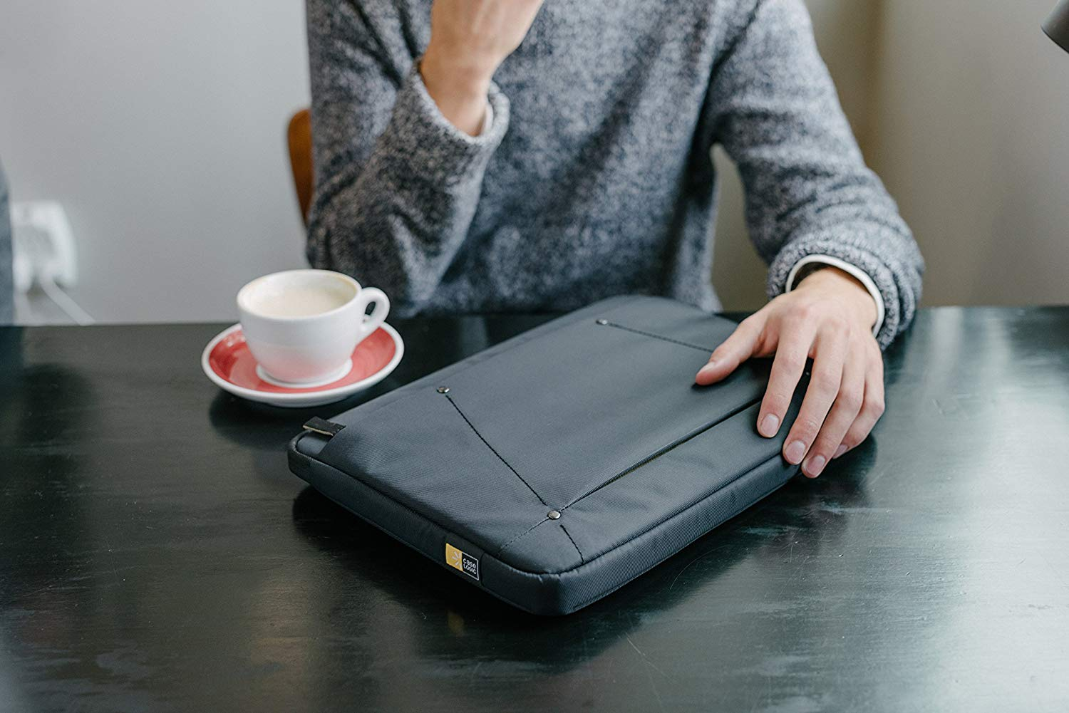 Egy személy egy laptop tokot, egy csésze kávét és egy csészealjat tart egy fekete asztalon.