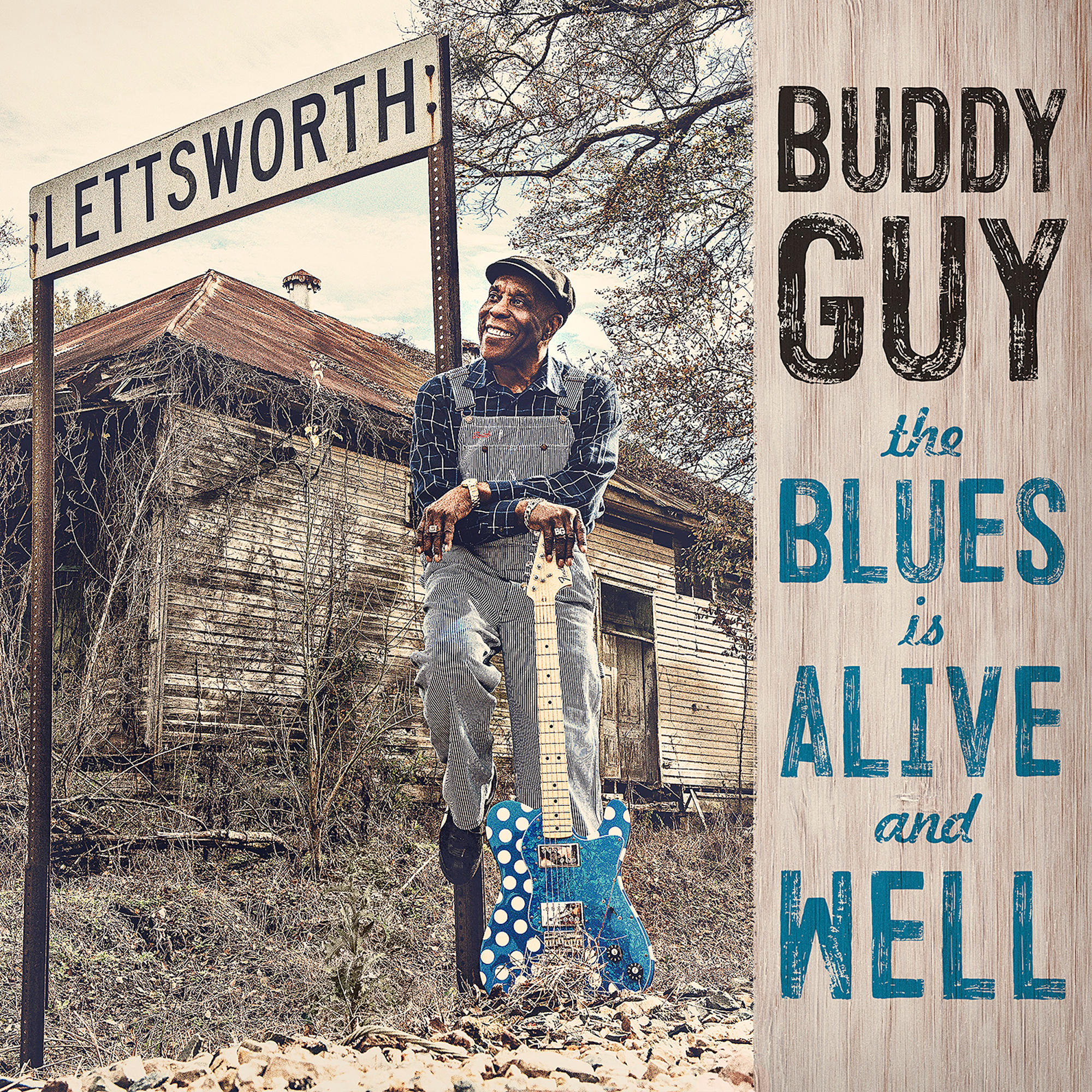 Buddy Guy mit einer blauen Gitarre, lehnt an einem Schild. Hintergrund ist ein baufälliges Gebäude und ein Holzschild.