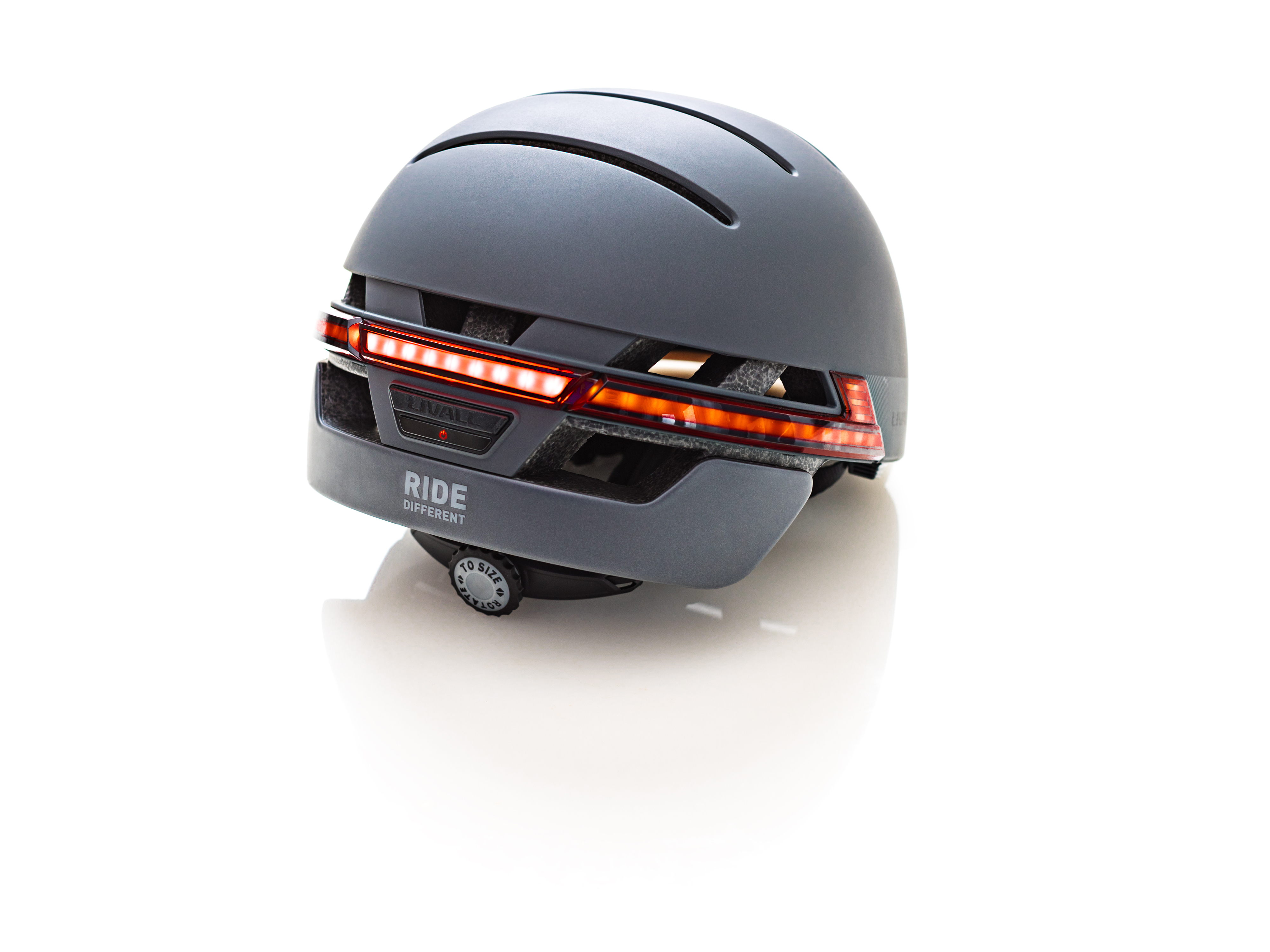 Grauer Fahrradhelm. Rote Lichter hinten. Text 'Ride Different'. Weißer Hintergrund.