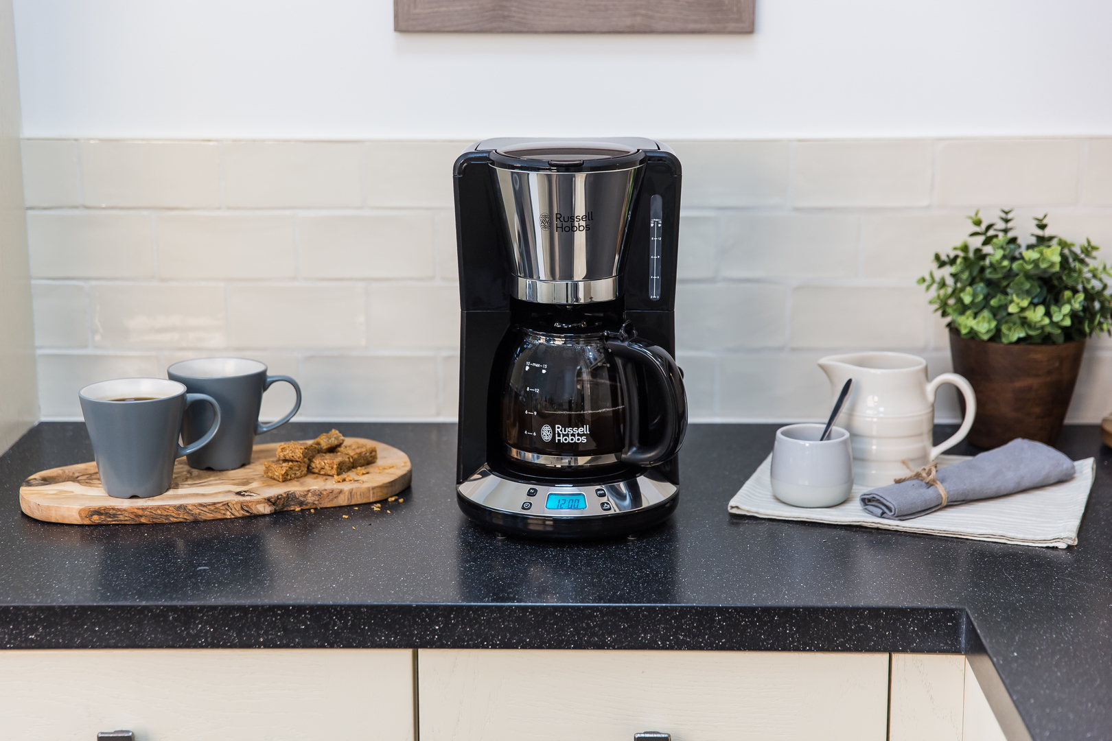Eine schwarze Kaffeemaschine auf einer Theke mit Tassen, Gebäck und einer Pflanze.