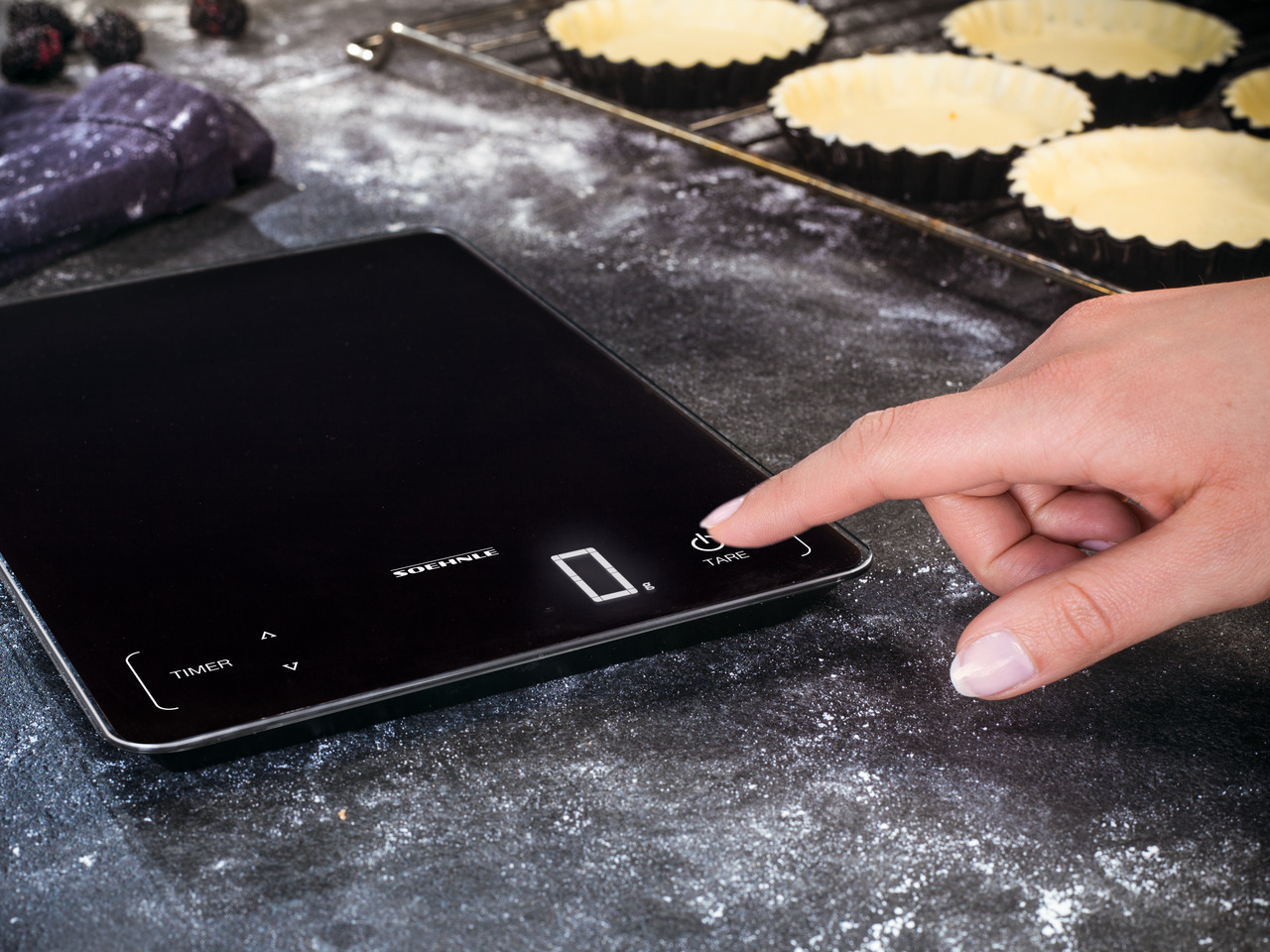 Eine schwarze digitale Küchenwaage mit einer Hand einer Frau, die die Taste 'TARE' drückt. Essen im Hintergrund.