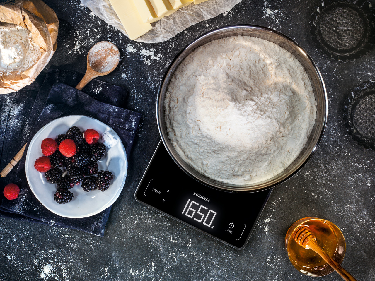 Waage mit Glasschüssel Mehl, Beeren, Butter und Honig.