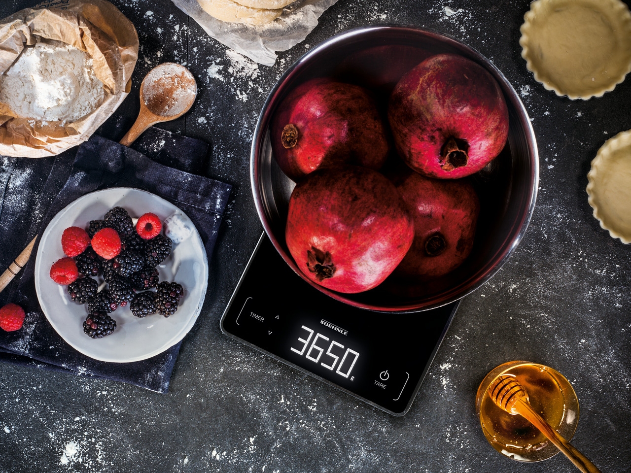Draufsicht auf eine Waage mit Granatäpfeln, Brombeeren und Honig. Backszene mit Mehl und Tortenböden.
