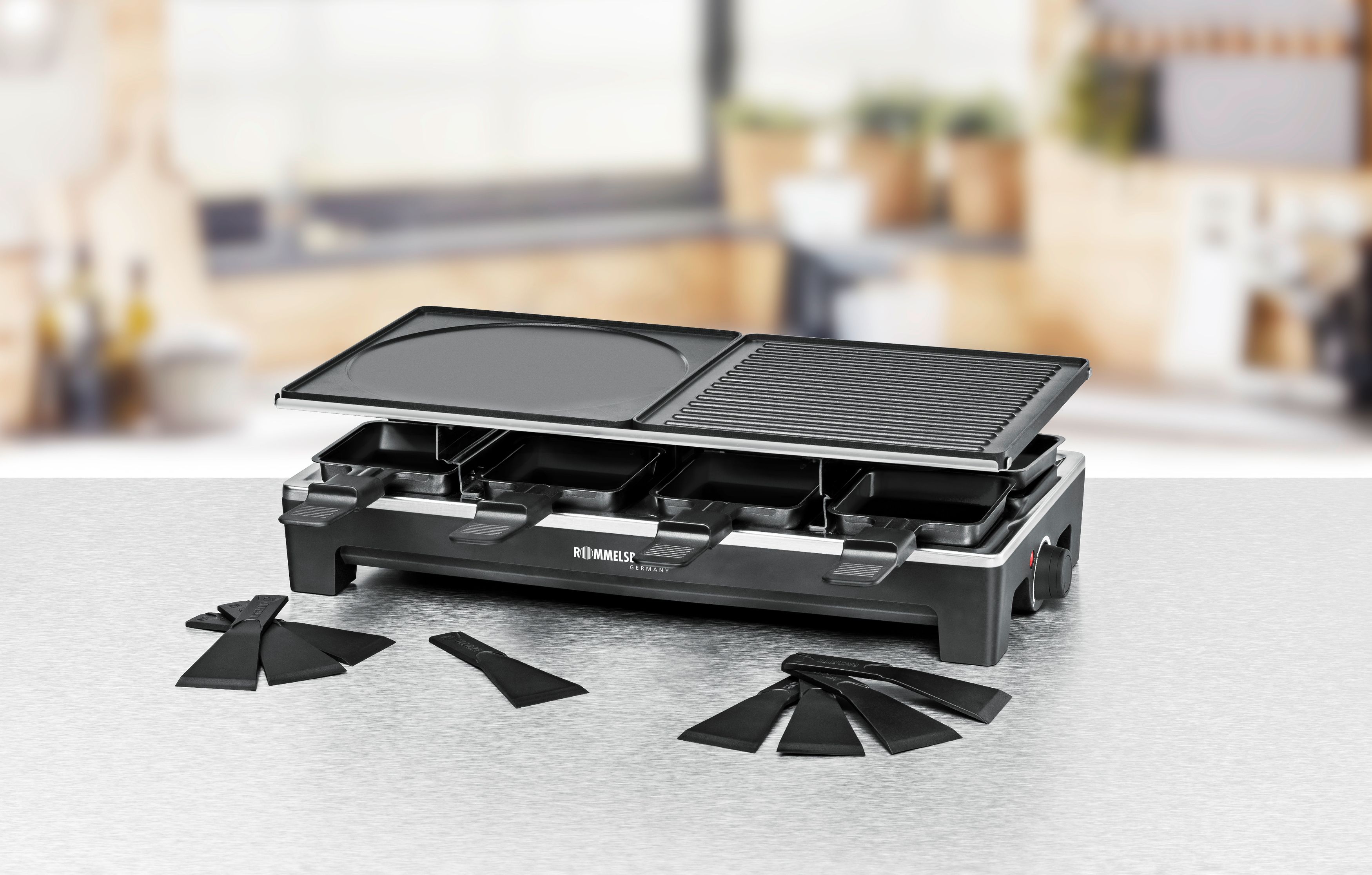 Ein Raclette-Grill mit offenem Deckel und Spateln, auf einer Theke mit verschwommenem Hintergrund.