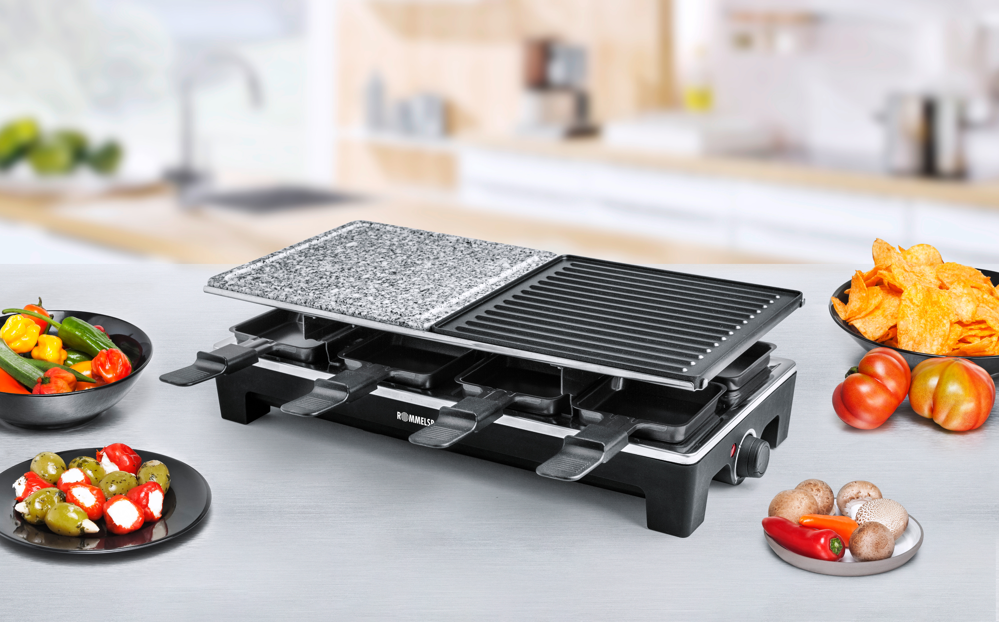 Ein Raclette-Grill mit Essen darauf, gebrauchsfertig. Teller mit Essen umgeben den Grill.