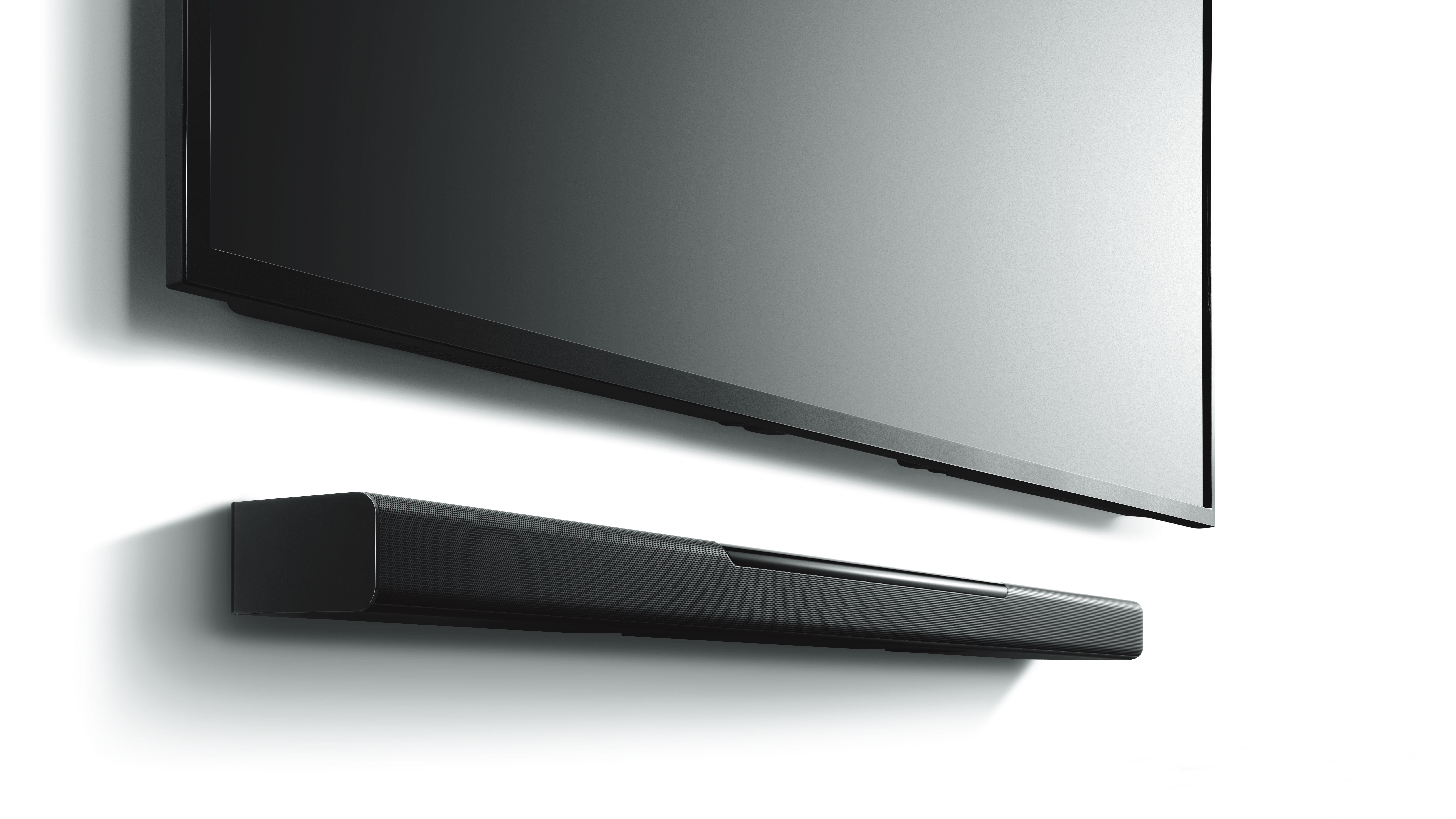 Eine schwarze Soundbar unter einem großen schwarzen Fernseher montiert. Fernseher und Soundbar sind an einer weißen Wand.