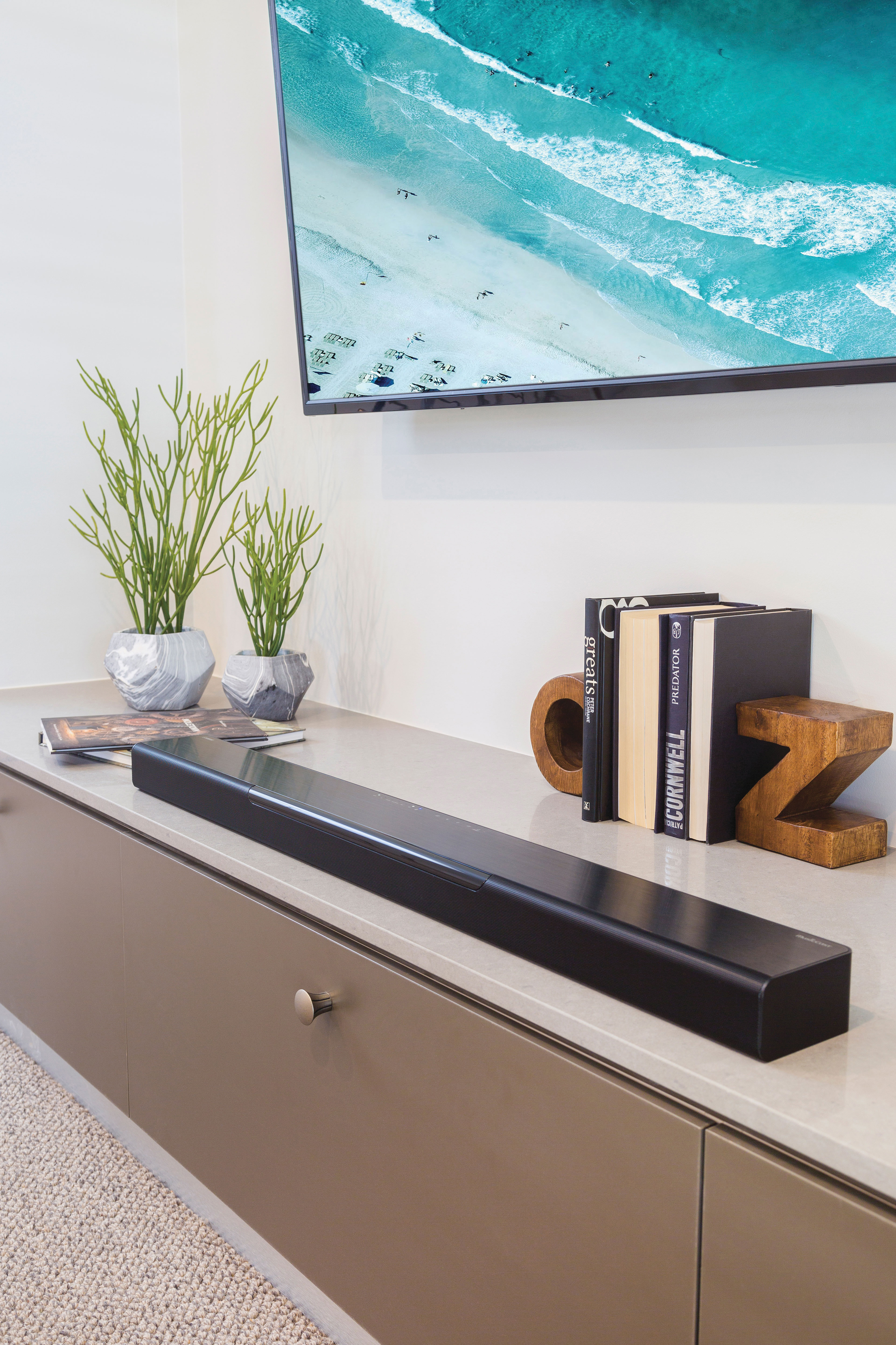 Eine schwarze Soundbar auf einem Schrank mit Pflanzen und Büchern. Ein Fernseher hängt darüber.