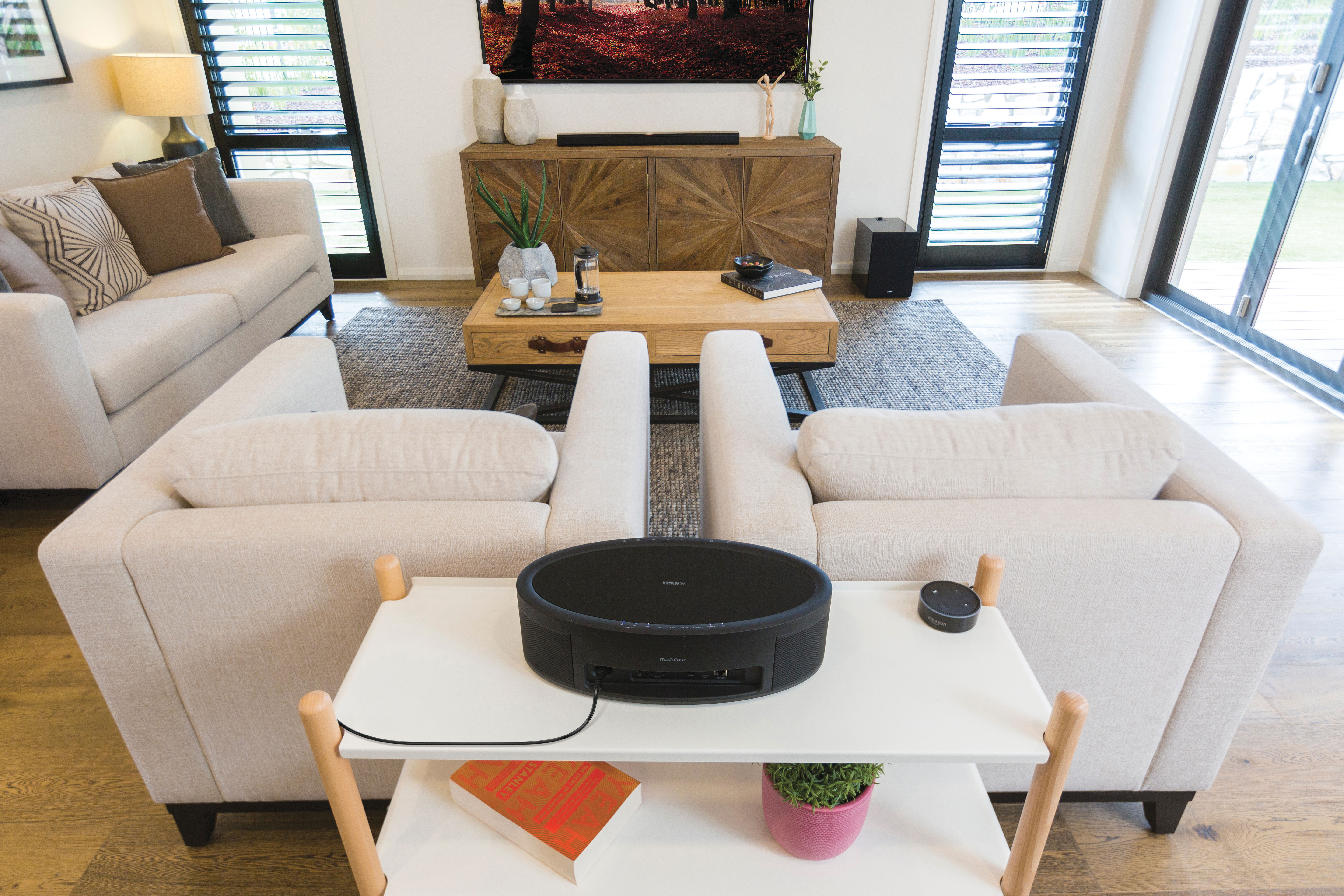 Wohnzimmer mit beigen Sofas, Holzmöbeln und einem Couchtisch. Auf einem weißen Beistelltisch steht ein Lautsprecher.