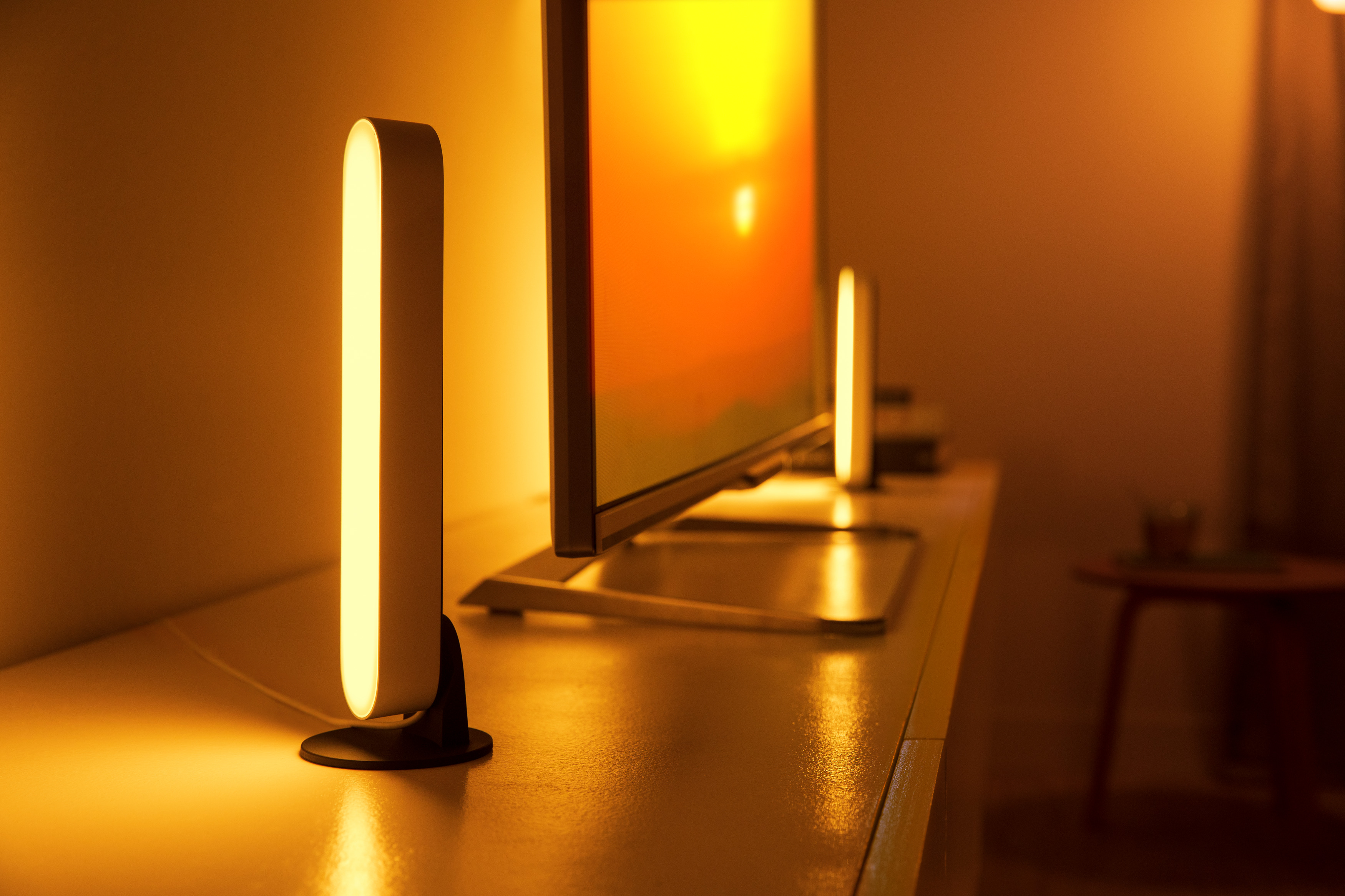 Zwei vertikale Lichter auf einem Tisch in der Nähe eines Fernsehers. Die Lichter sind weiß und der Fernseher ist an.