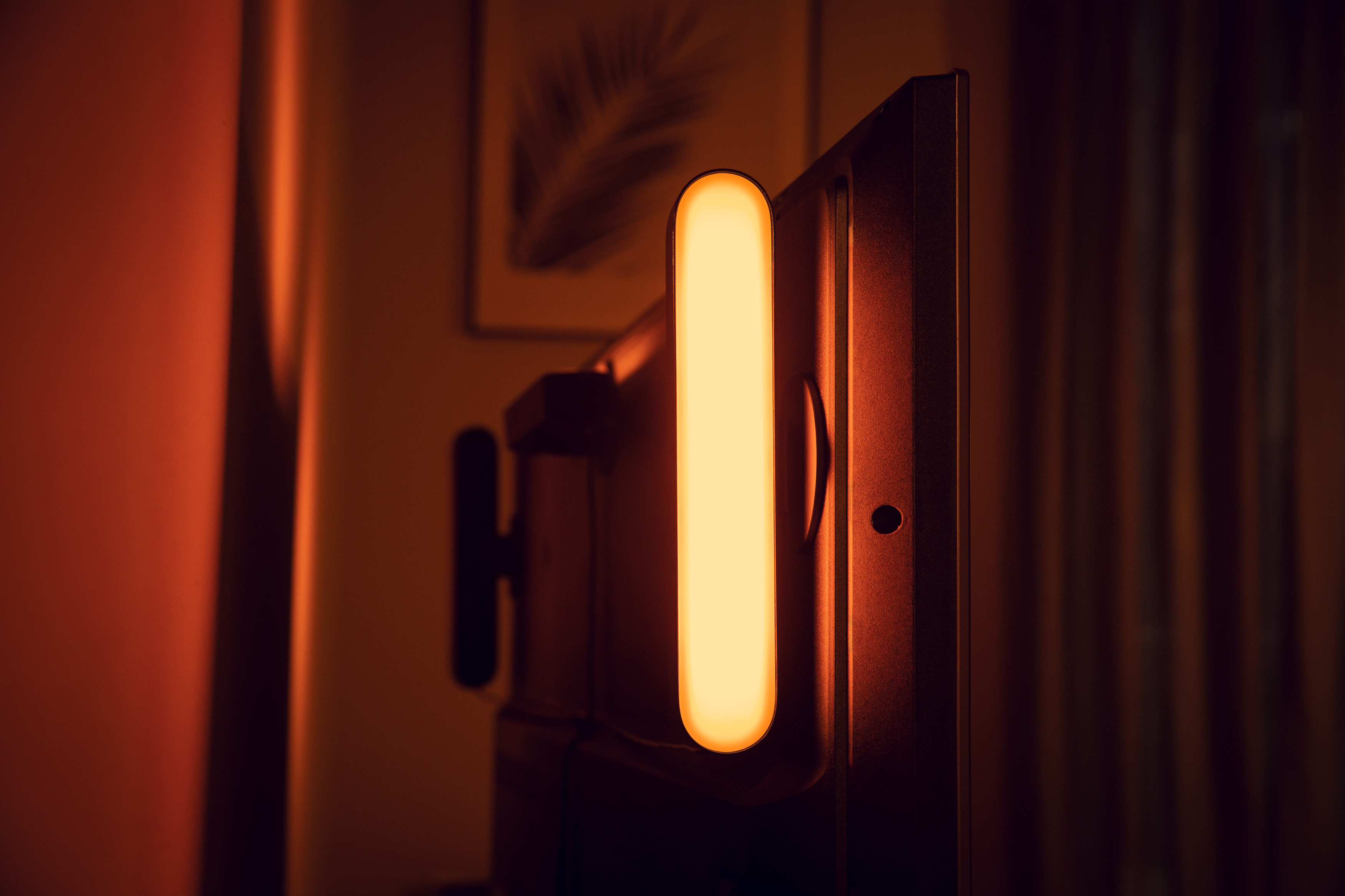 Eine lange, beleuchtete Lichtleiste neben einem dunklen Fernsehbildschirm, vor einer orangefarbenen Wand.