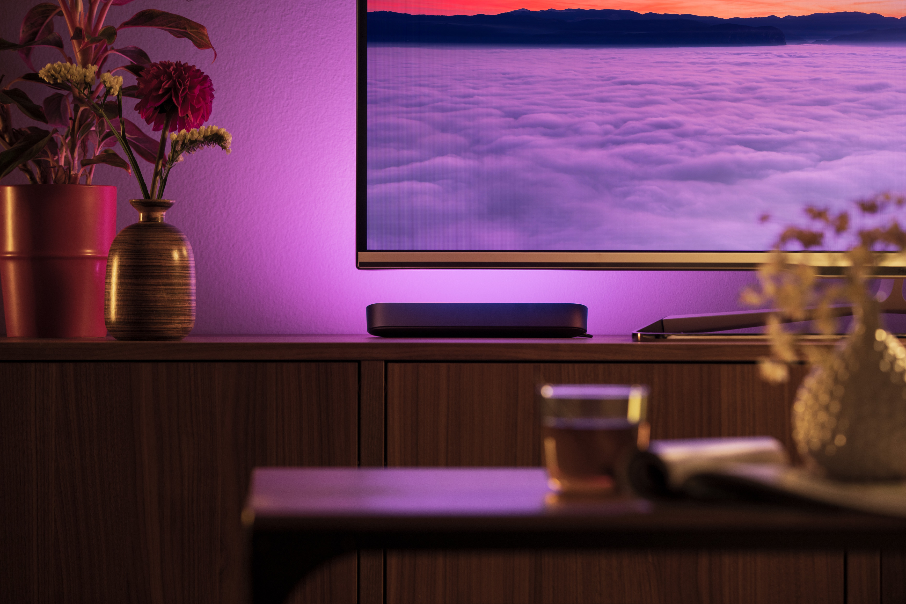 Wohnzimmerszene mit Fernseher, Soundbar und Schrank. Dekorative Pflanzen und lila Beleuchtung vorhanden.