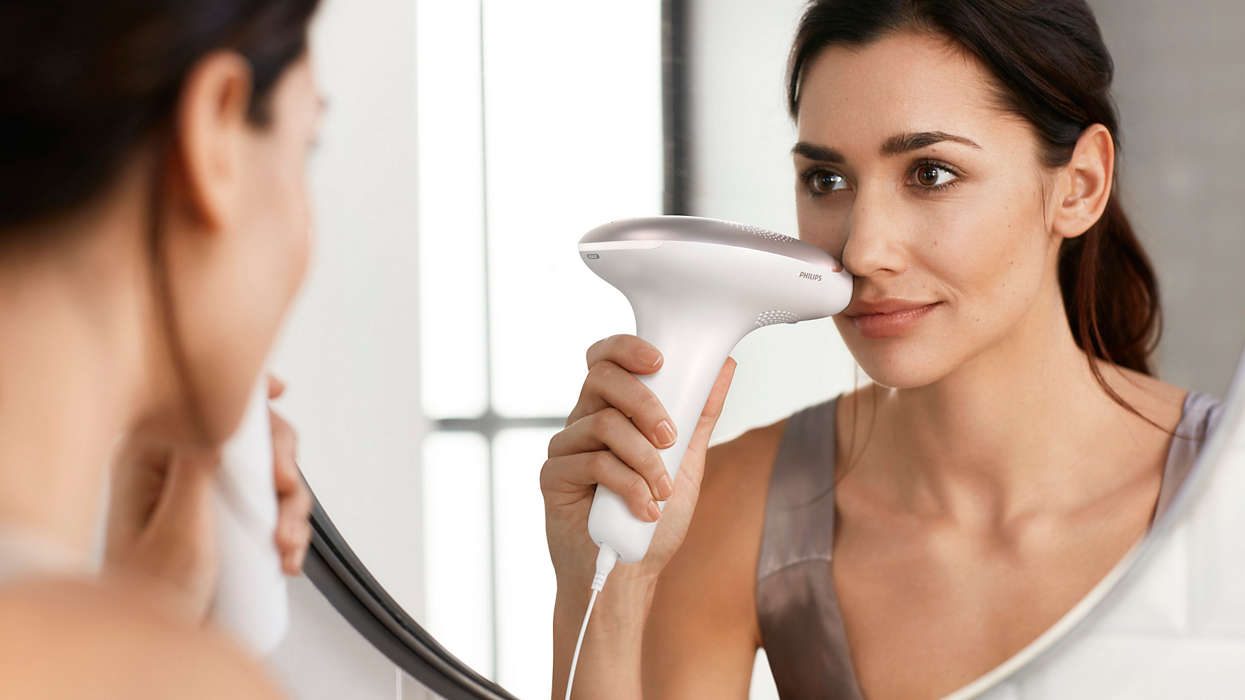A woman looking in a mirror using a facial hair removal device on her face.