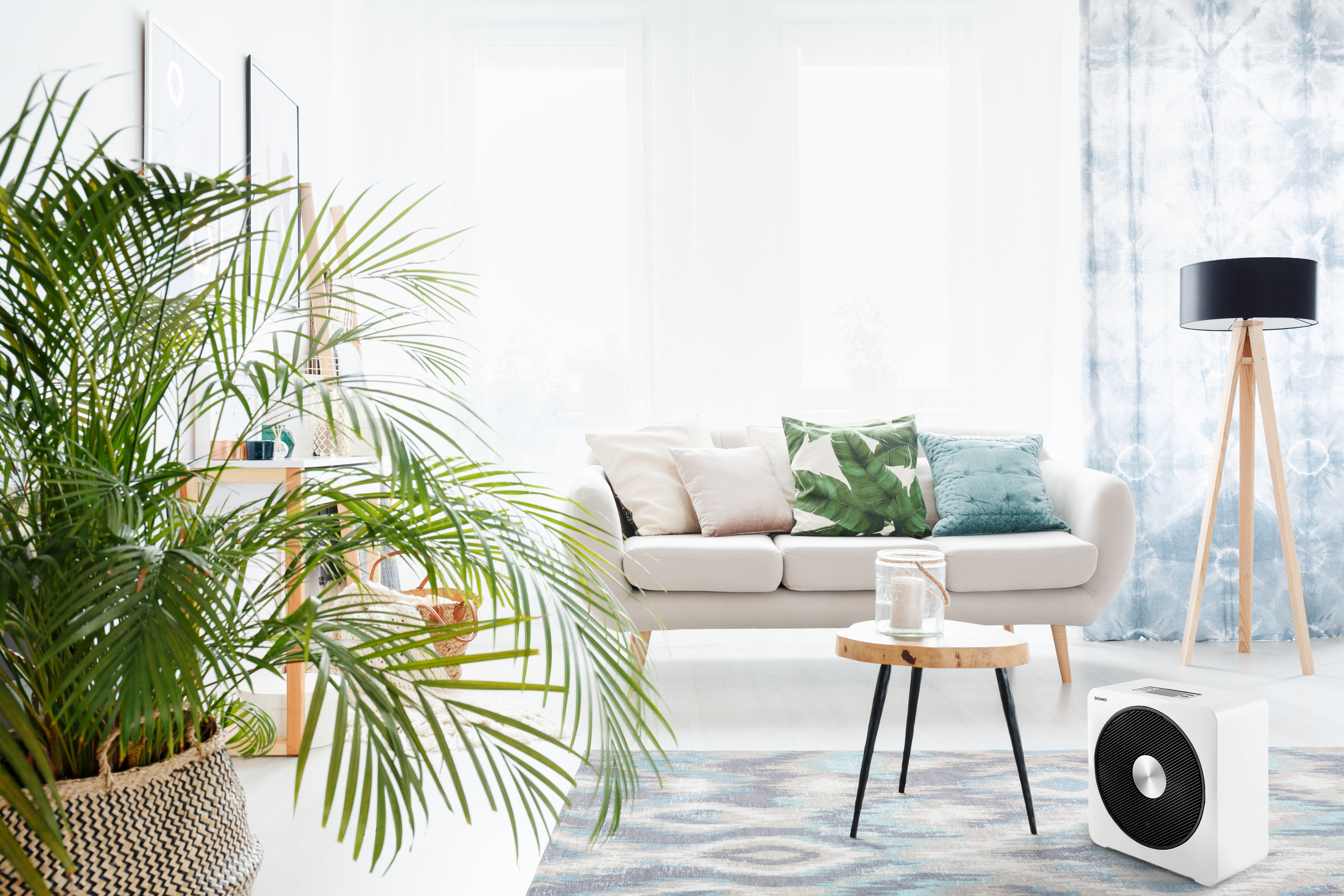 A living room with a white sofa, plants, and a wooden floor lamp. A white heater is in the foreground.