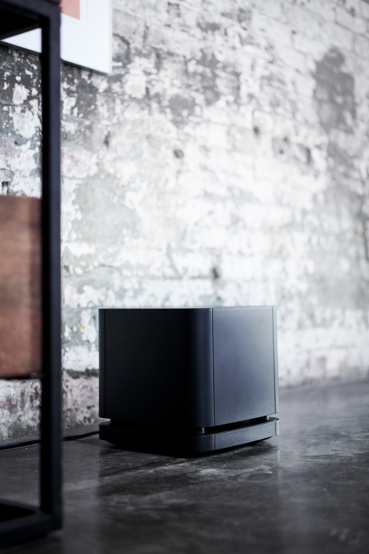 Schwarzer Subwoofer auf dem Boden neben einem Metallrahmen und einer Backsteinmauer.
