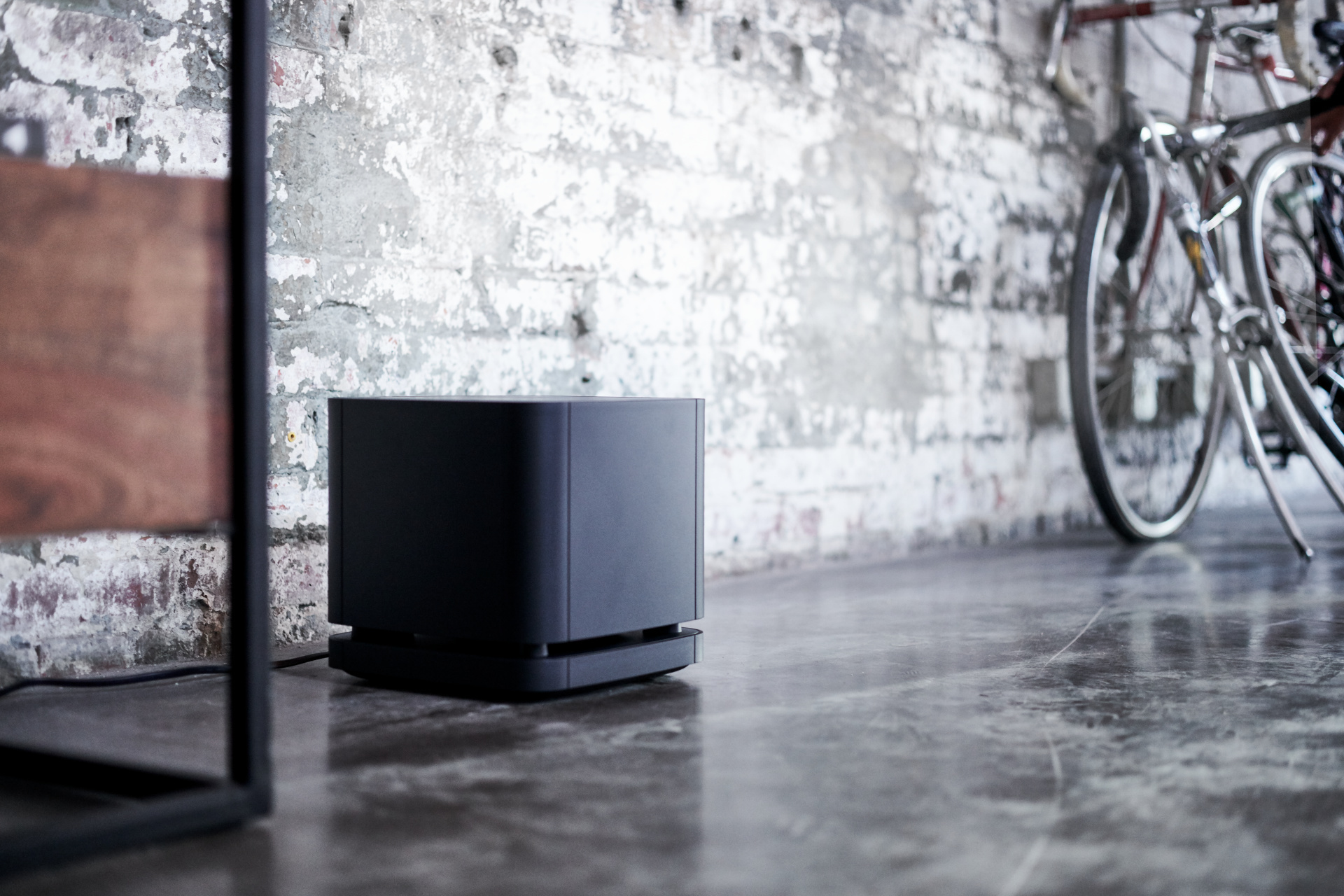 Ein schwarzer Subwoofer auf einem Betonboden. Eine Ziegelmauer und Fahrräder befinden sich im Hintergrund.