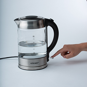 Ein elektrischer Glaskessel mit Wasser. Eine Hand berührt den Ein-/Ausschalter.
