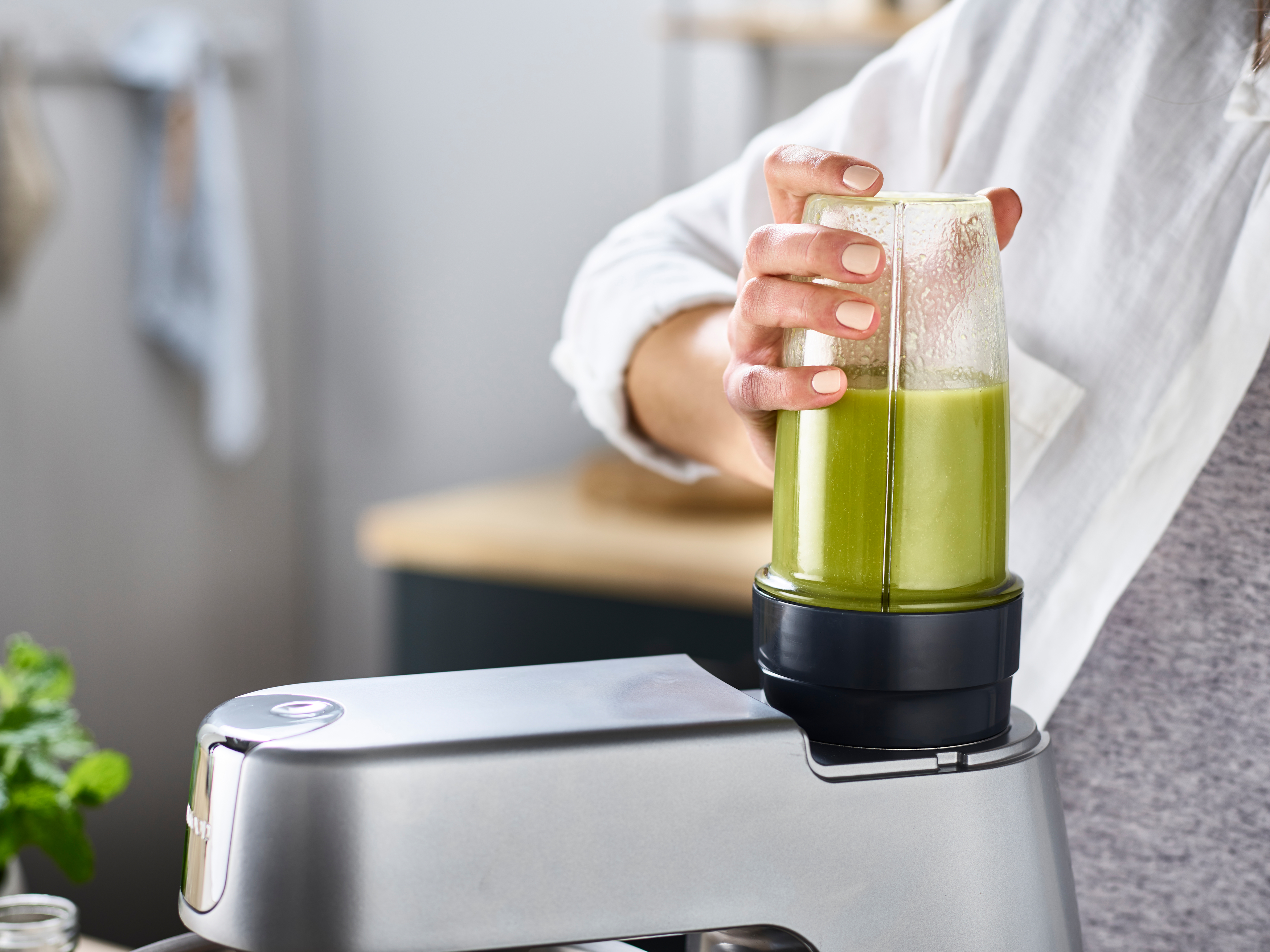 Eine Frau hält einen Mixer mit grüner Flüssigkeit, befestigt an einer Küchenmaschine.