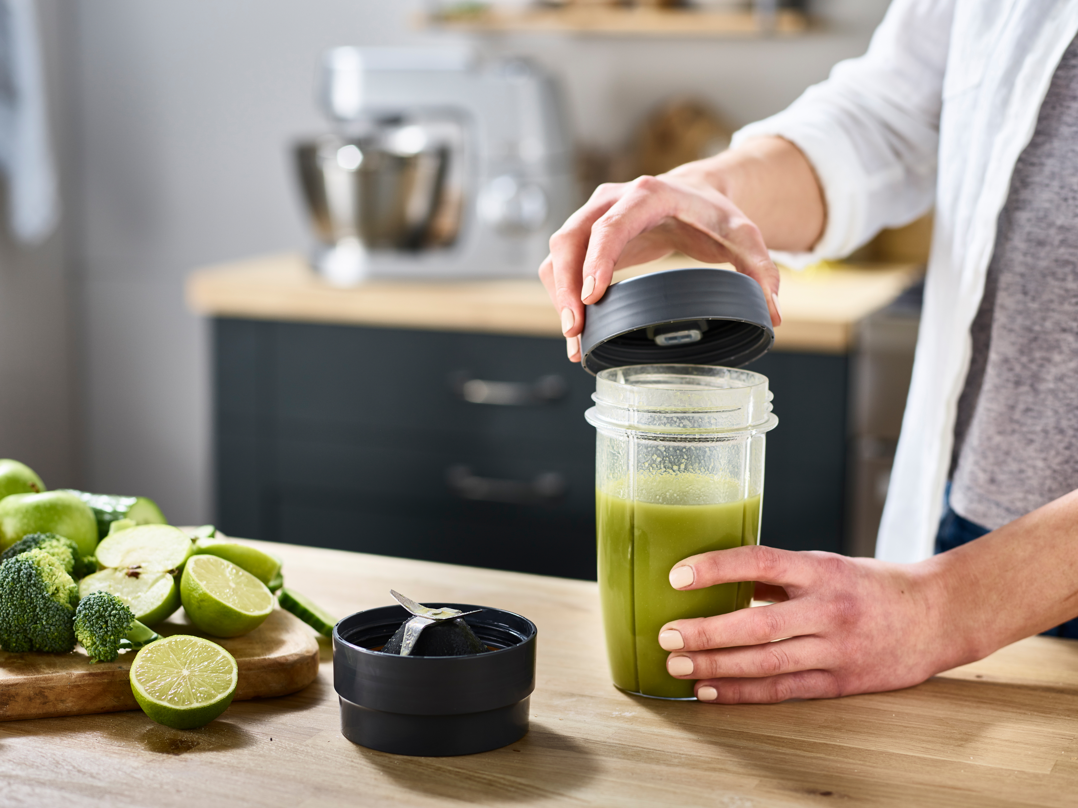 Frauenhände öffnen Mixer. Grüner Smoothie sichtbar. Geschnittene Limetten und Gemüse liegen auf dem Tisch.