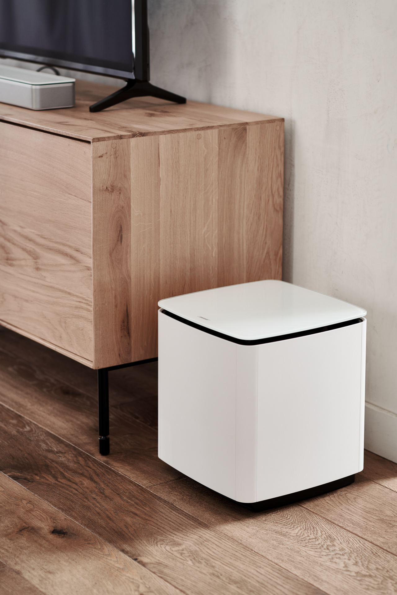 Ein weißer Subwoofer auf einem Holzboden, neben einem Holzschrank und einem Fernseher.