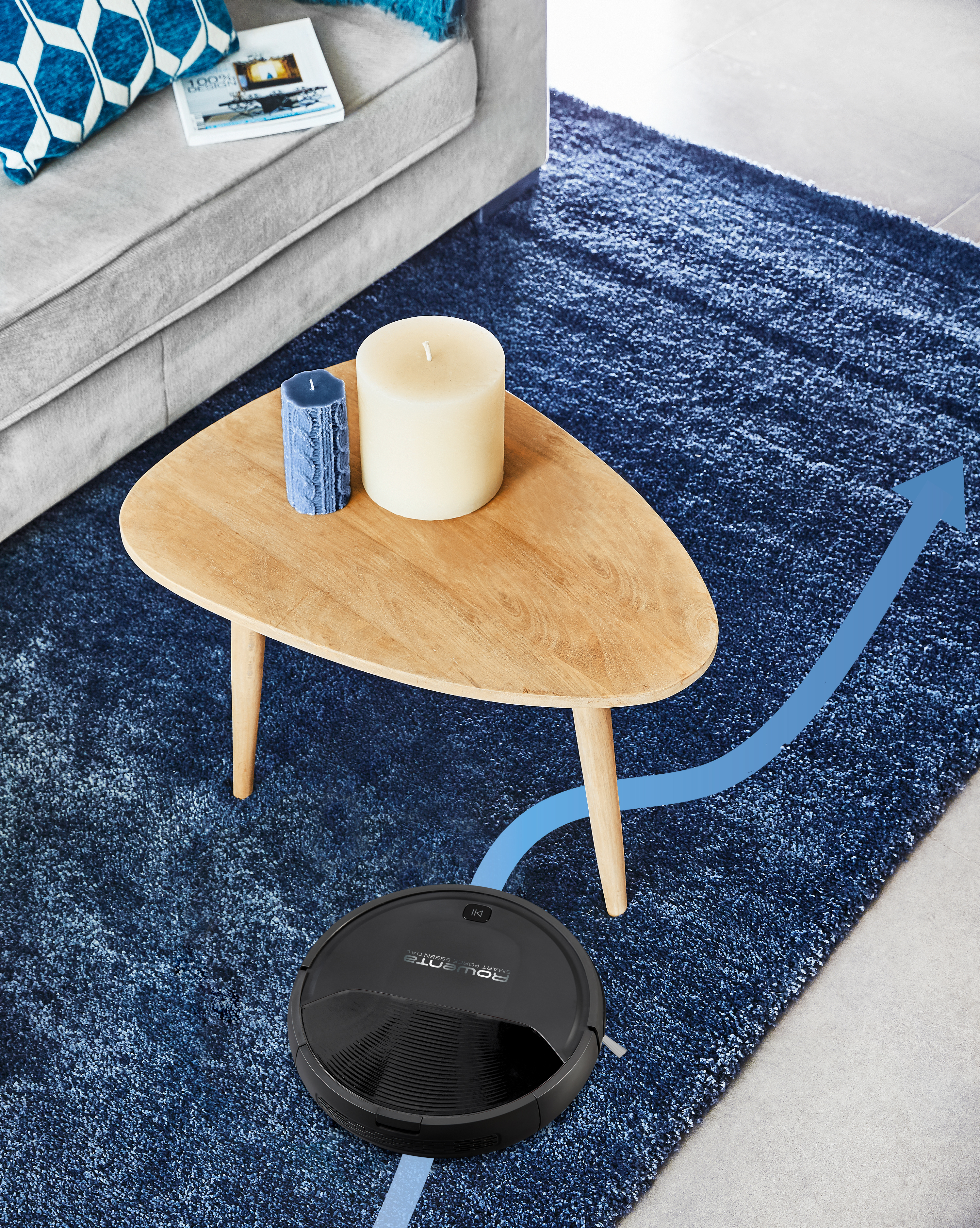 Ein Roboterstaubsauger reinigt einen blauen Teppich. Ein Couchtisch mit Kerzen befindet sich darüber. Im Hintergrund befindet sich ein Sofa mit Kissen.