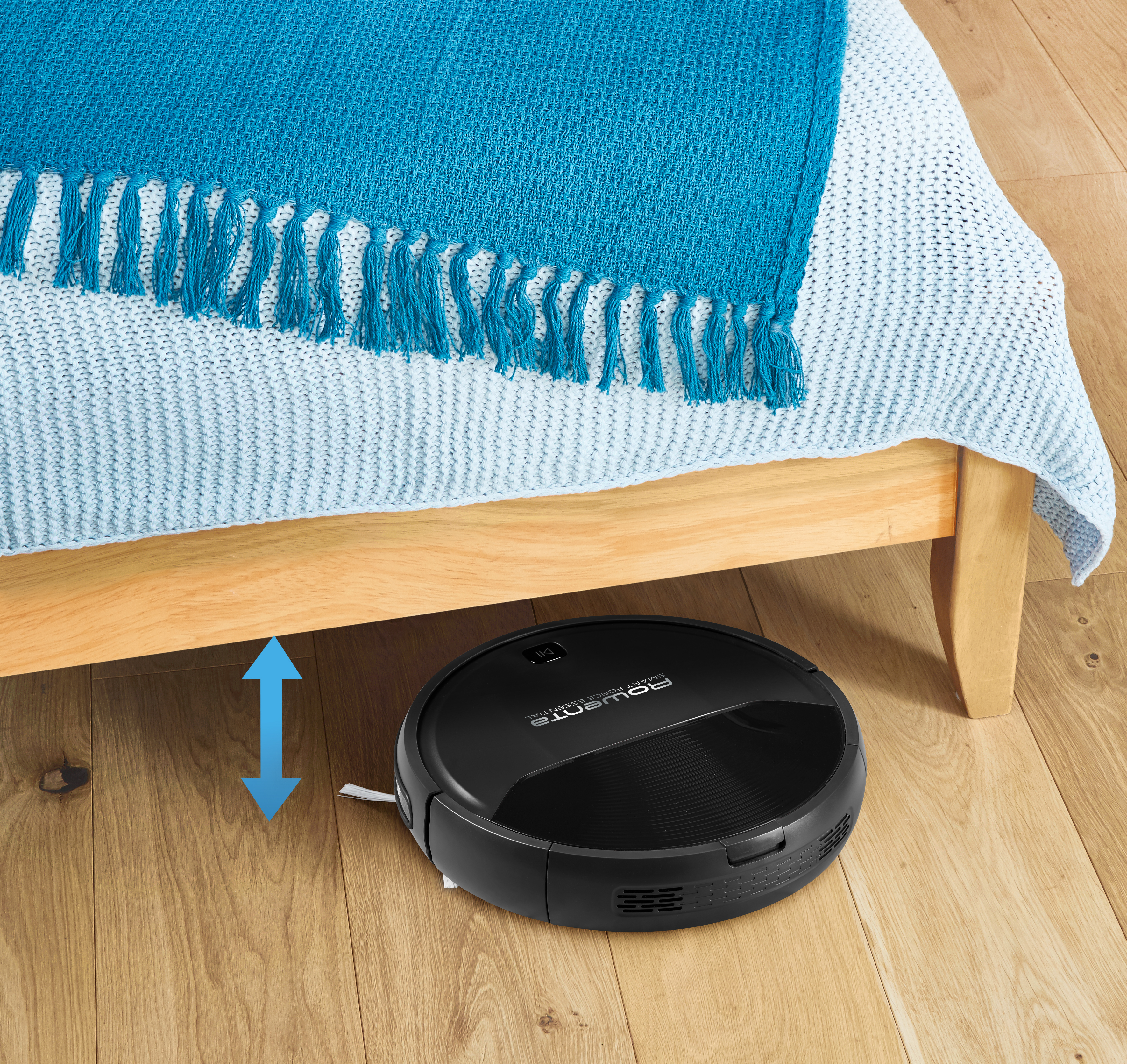 Schwarzer Staubsaugerroboter unter einem Bett mit blauer Decke. Holzboden.