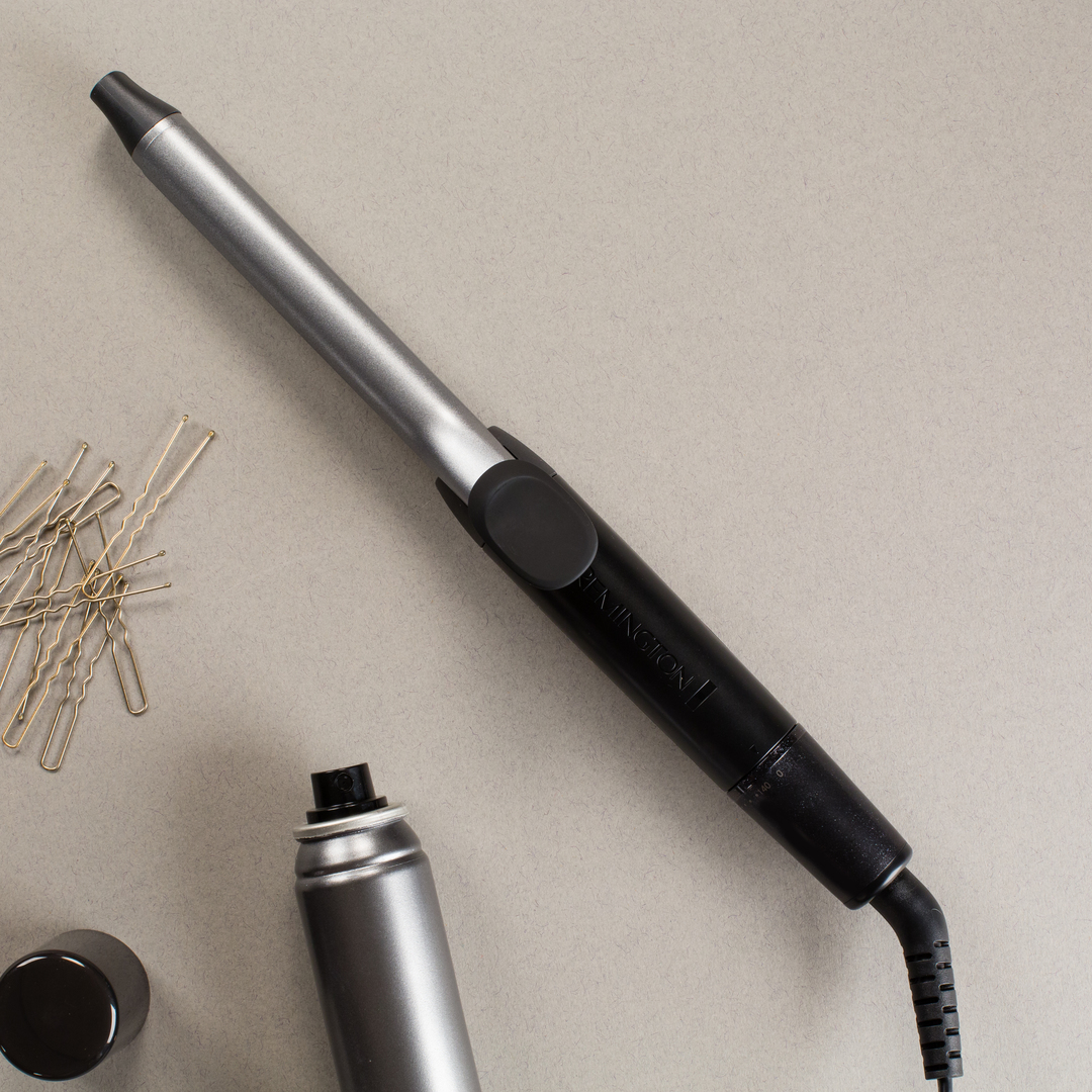 Hair curler and accessories on a grey surface. The curler is silver and black, with a spray and pins.