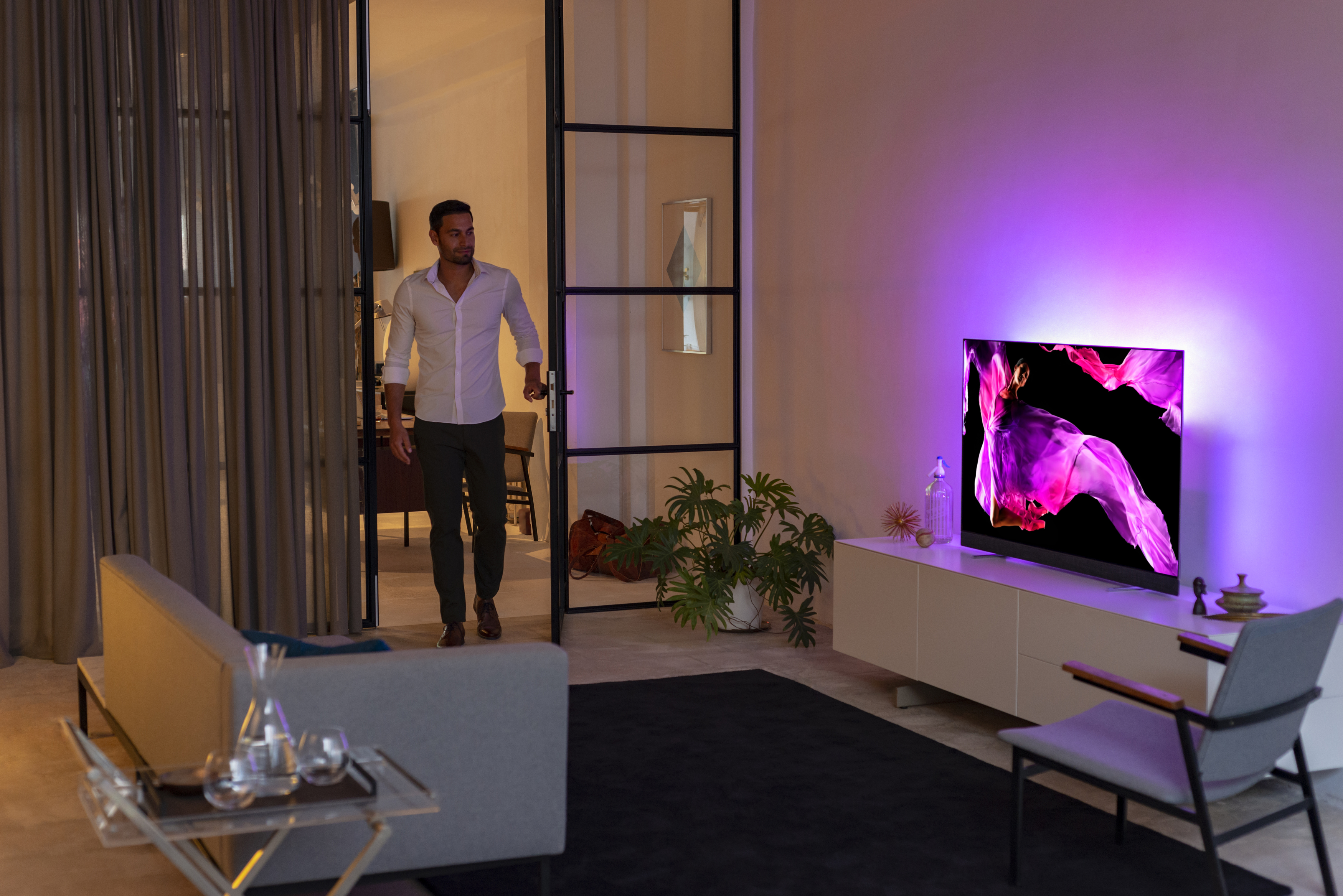 Ein Mann betritt durch eine Tür ein Wohnzimmer mit einem Fernseher, der ein farbenfrohes Bild zeigt.