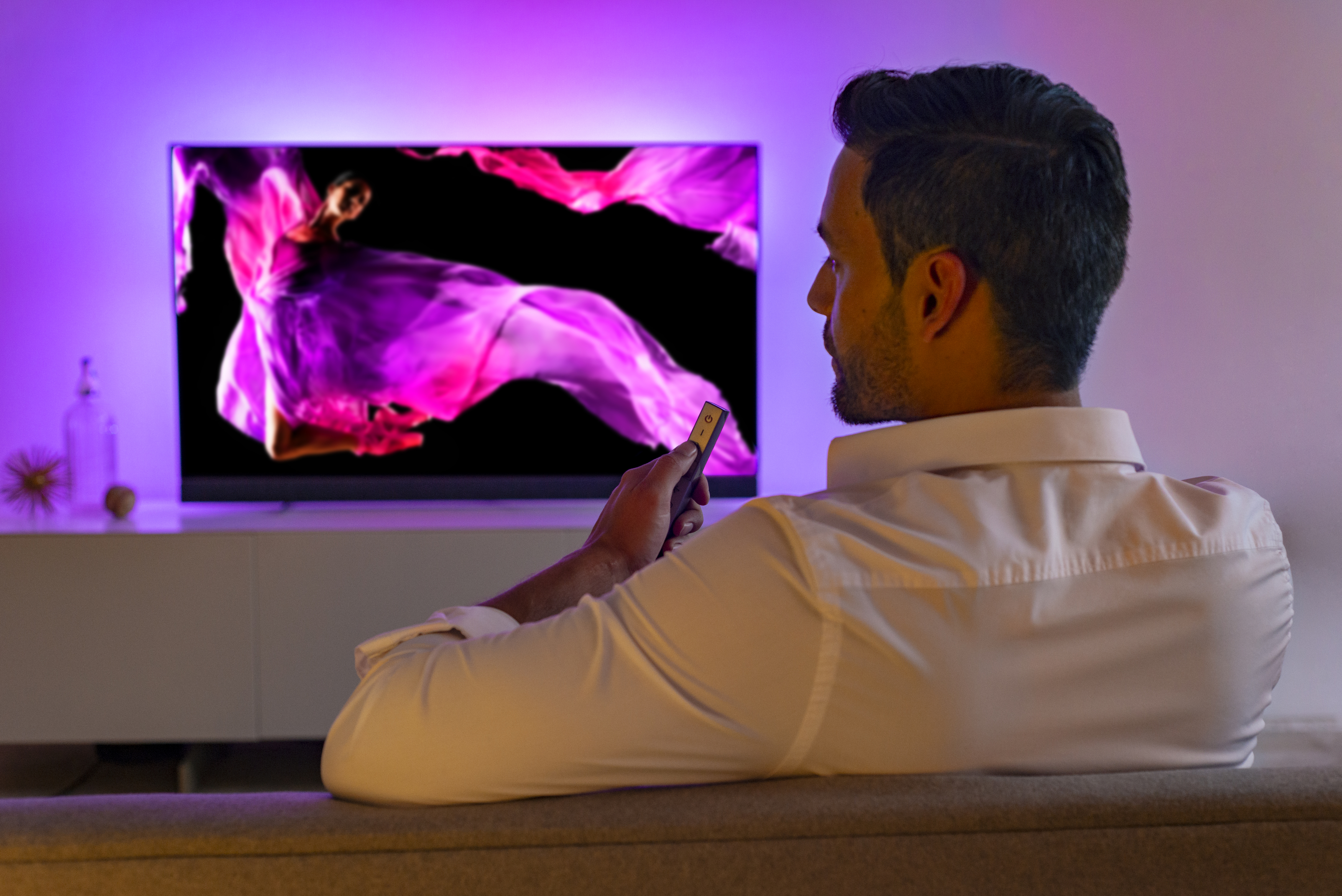 Mann sieht fern. Der Fernseher zeigt eine Frau in einem rosa Kleid vor schwarzem Hintergrund.