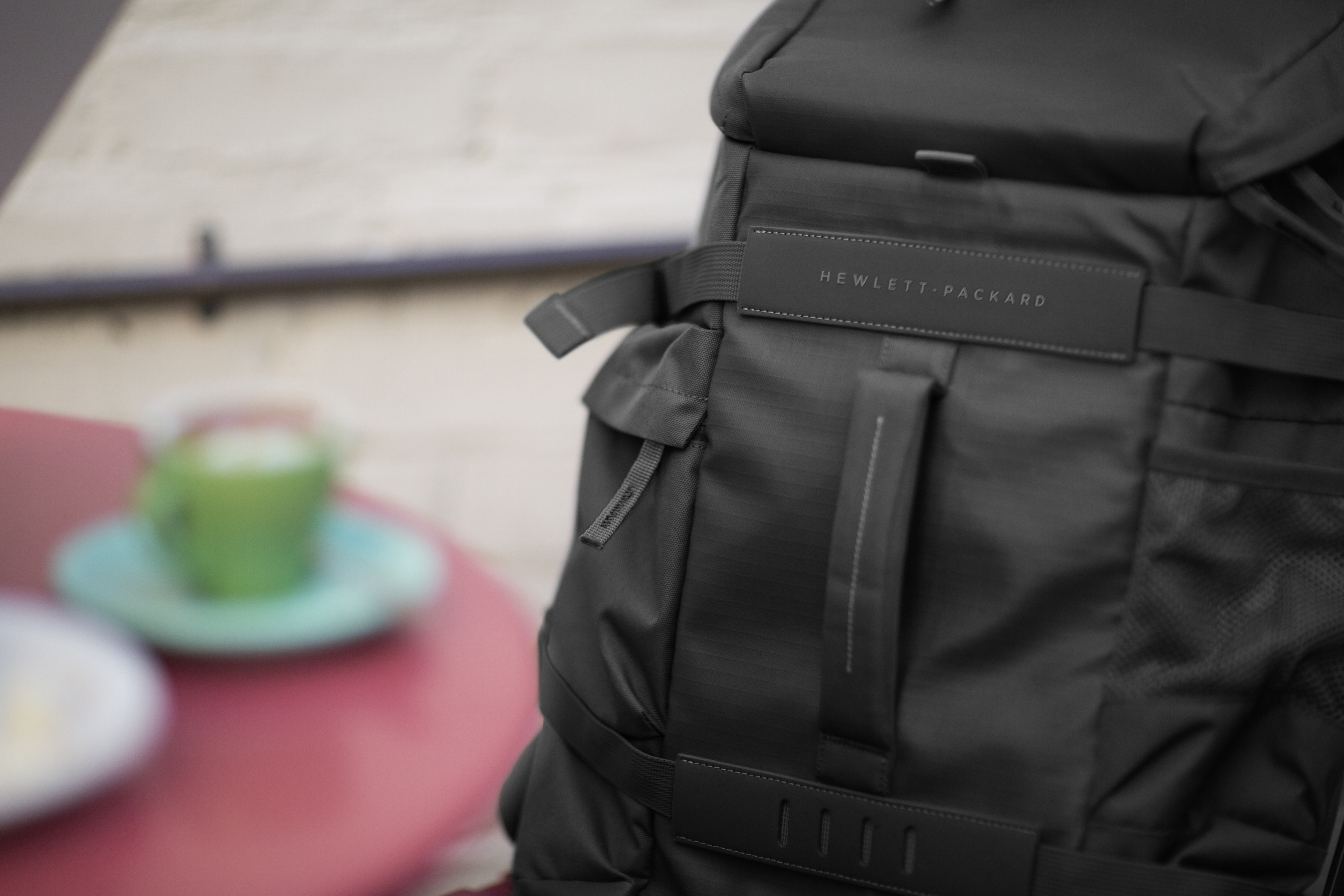 Siyah sırt çantası, Hewlett-Packard etiketi, bulanık bir kafe ortamında. Yakınlarda yeşil bir kupa.